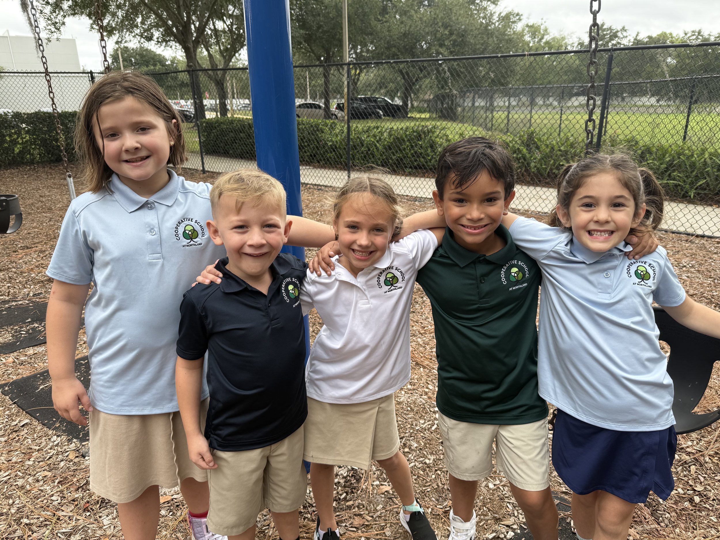 A group of five children smiling and standing close together outdoors at a playground. They are wearing school uniforms with a logo that says "Cooperative School at Northland." There is a fence, trees, and parked cars in the background.