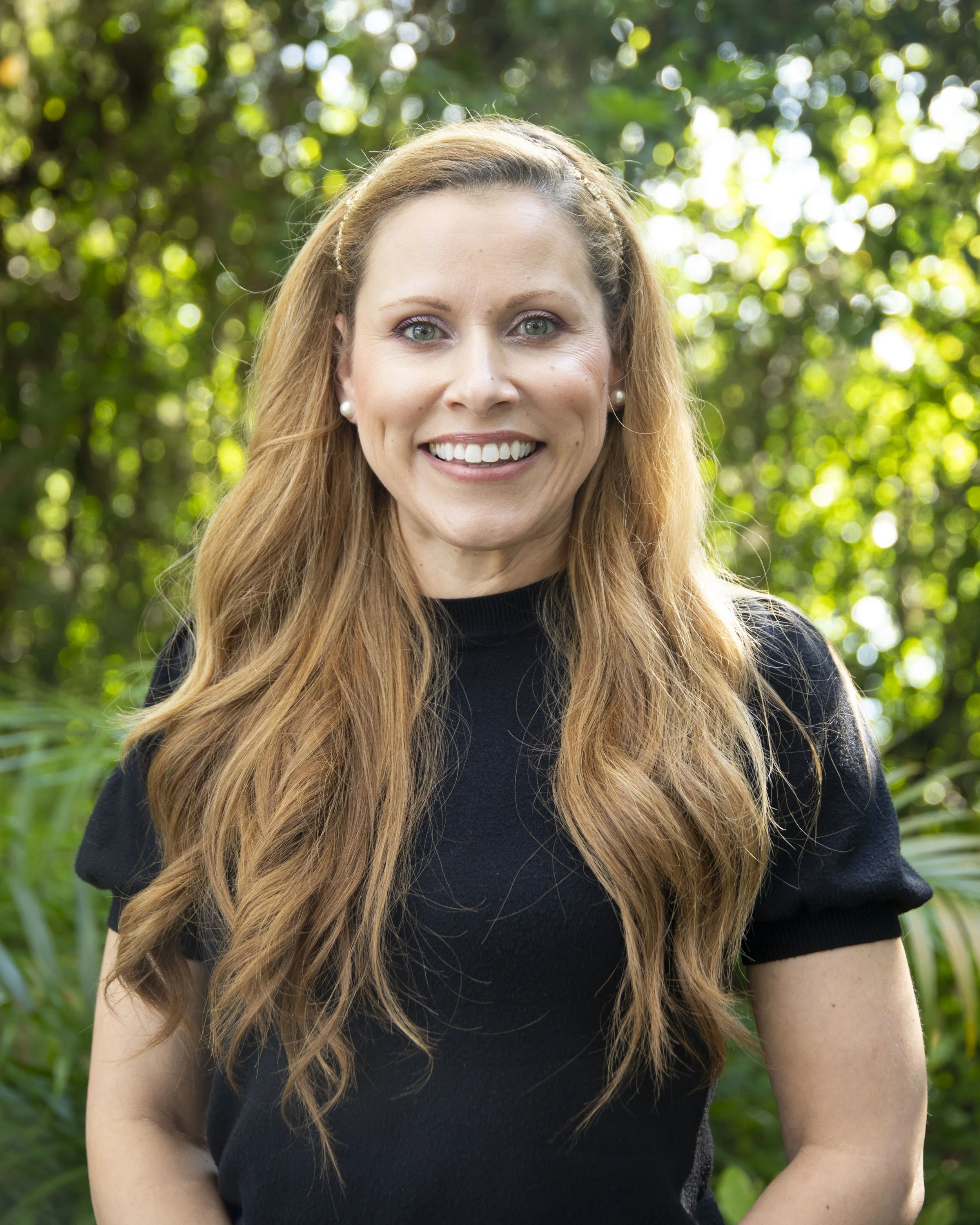 Close-up of a woman with long wavy red hair, wearing pearl earrings and a black shirt, smiling outdoors with trees and green foliage in the background.
