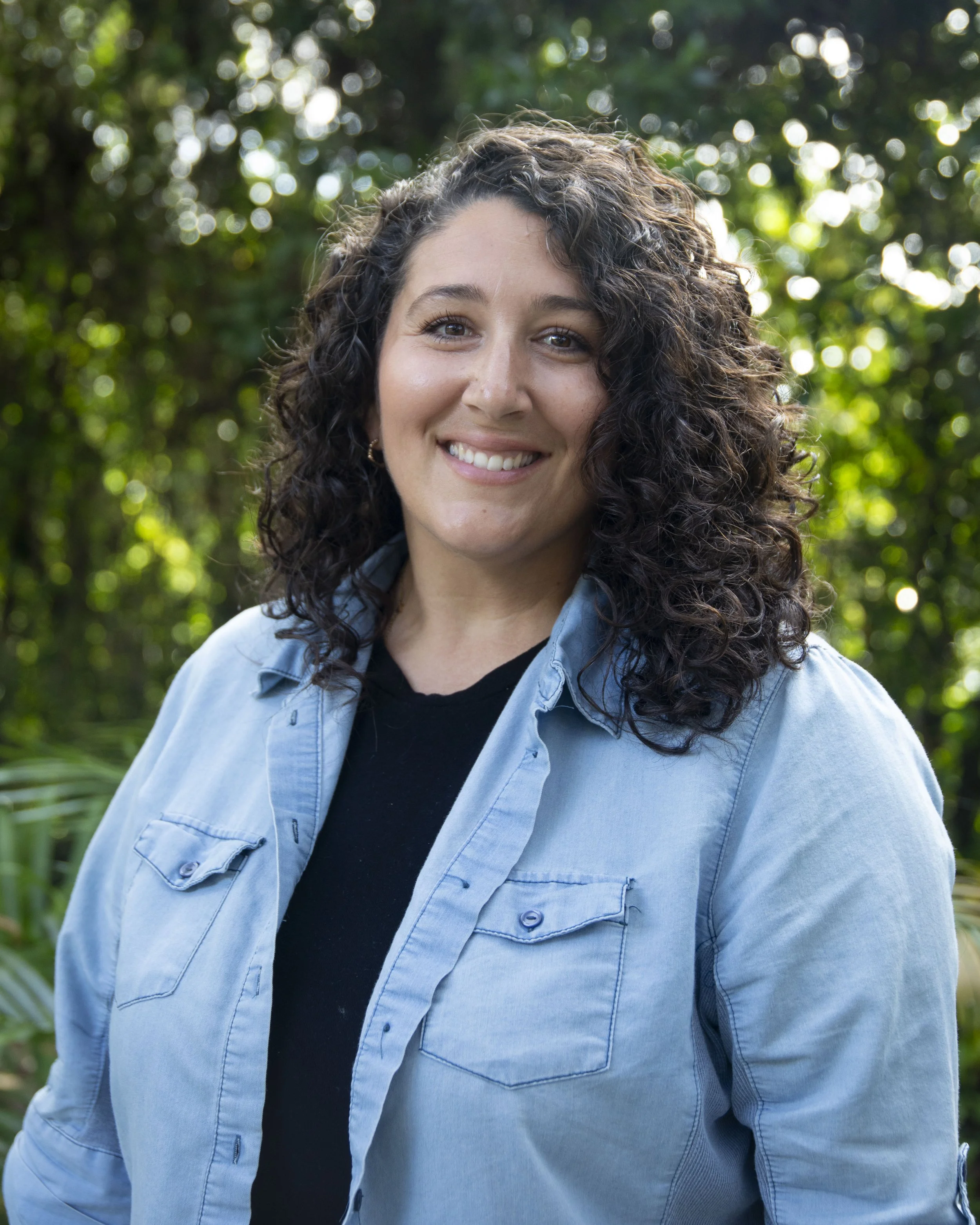 A young woman with curly dark hair smiling outdoors with green foliage in the background.