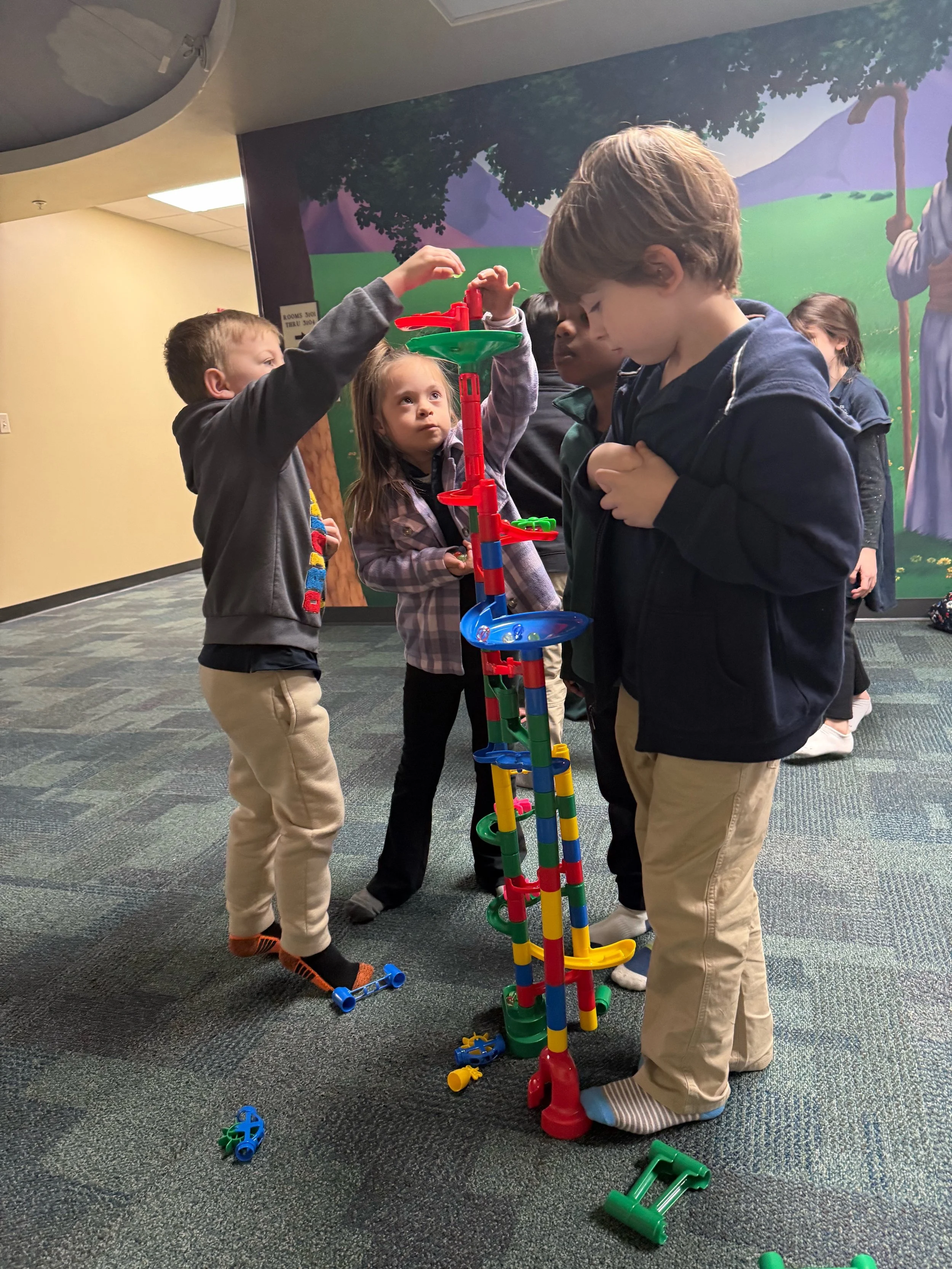 Children playing with a colorful marble run toy indoors with a painted mural in the background.
