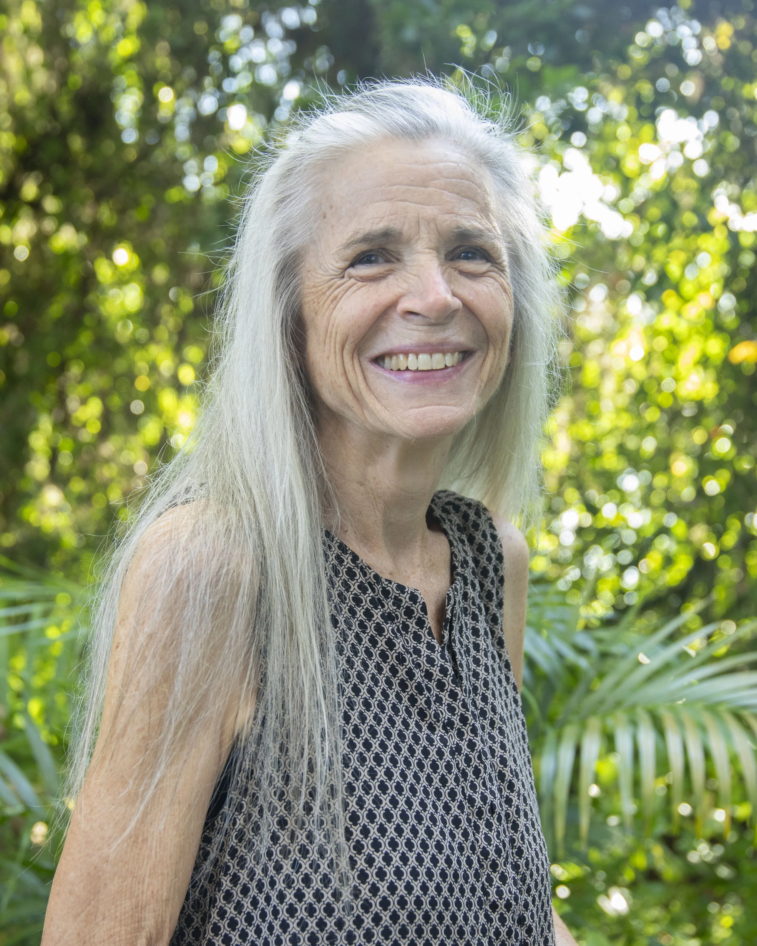 Close-up of an elderly woman with long silver hair smiling outdoors amidst green foliage.
