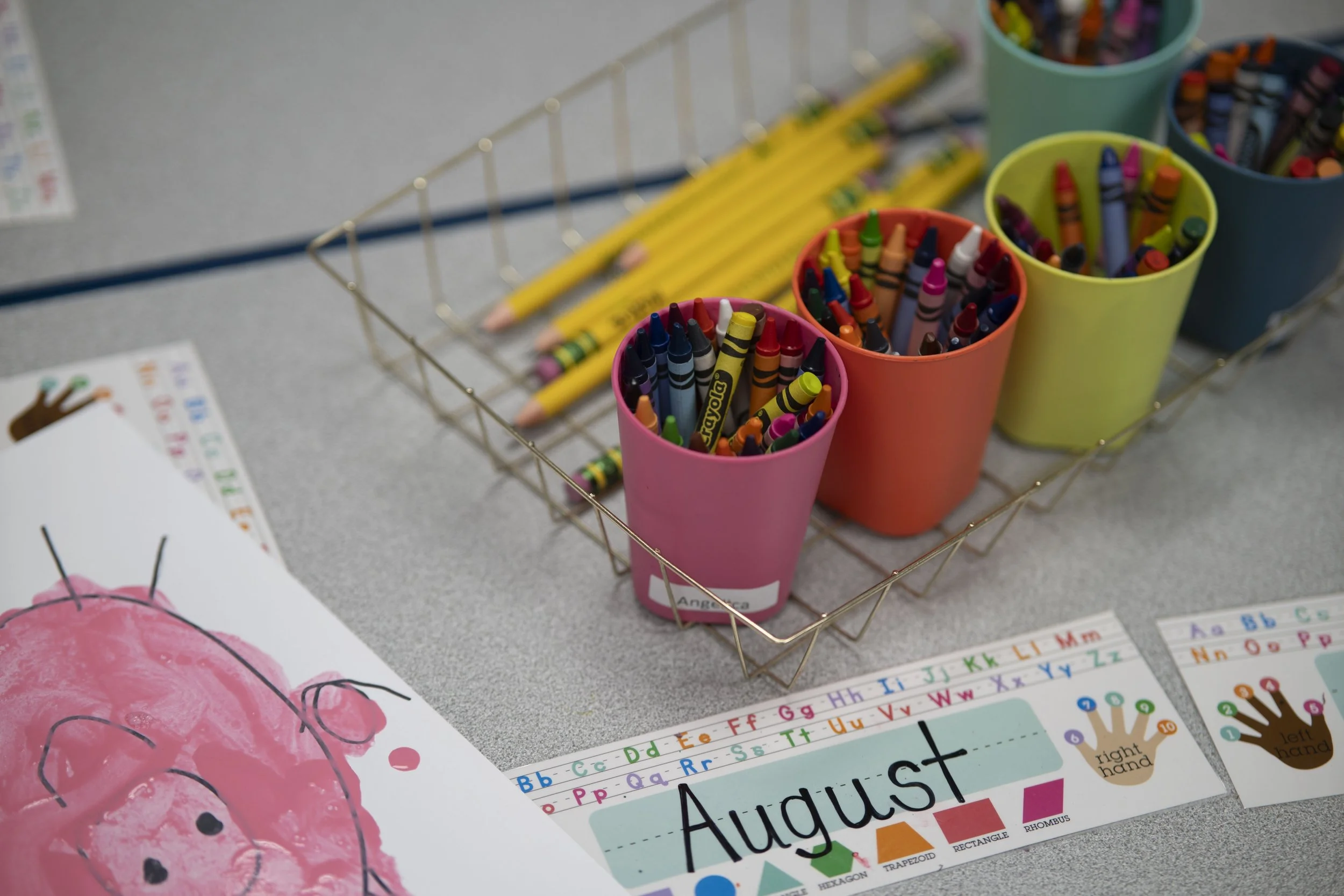 Colorful containers filled with crayons and pencils on a desk in a classroom, with an educational poster for the month of August visible in the foreground.