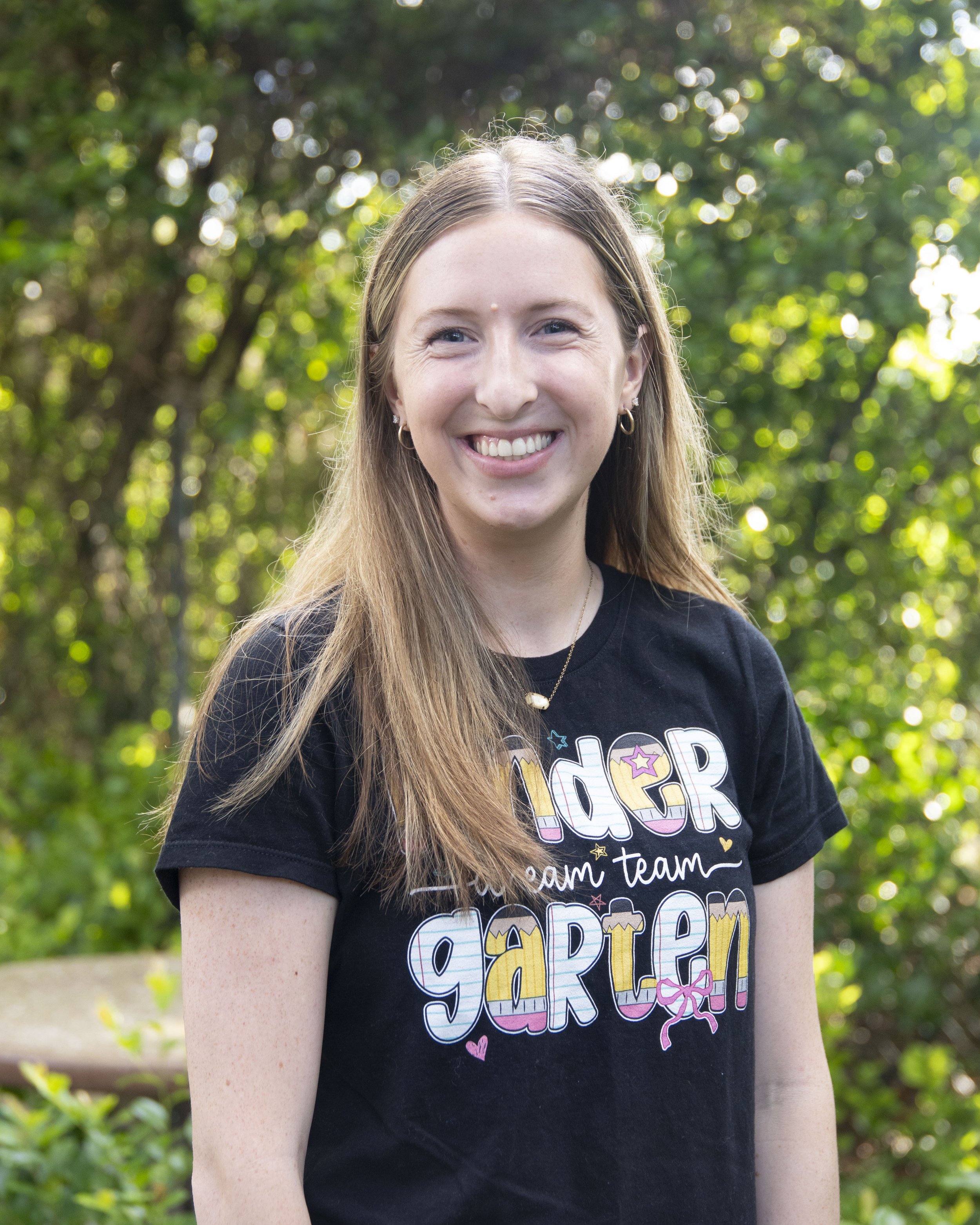 A young woman with long, light brown hair and blue eyes smiling outdoors, wearing a black t-shirt with colorful text and graphics, in front of a background of green trees and sunlight.