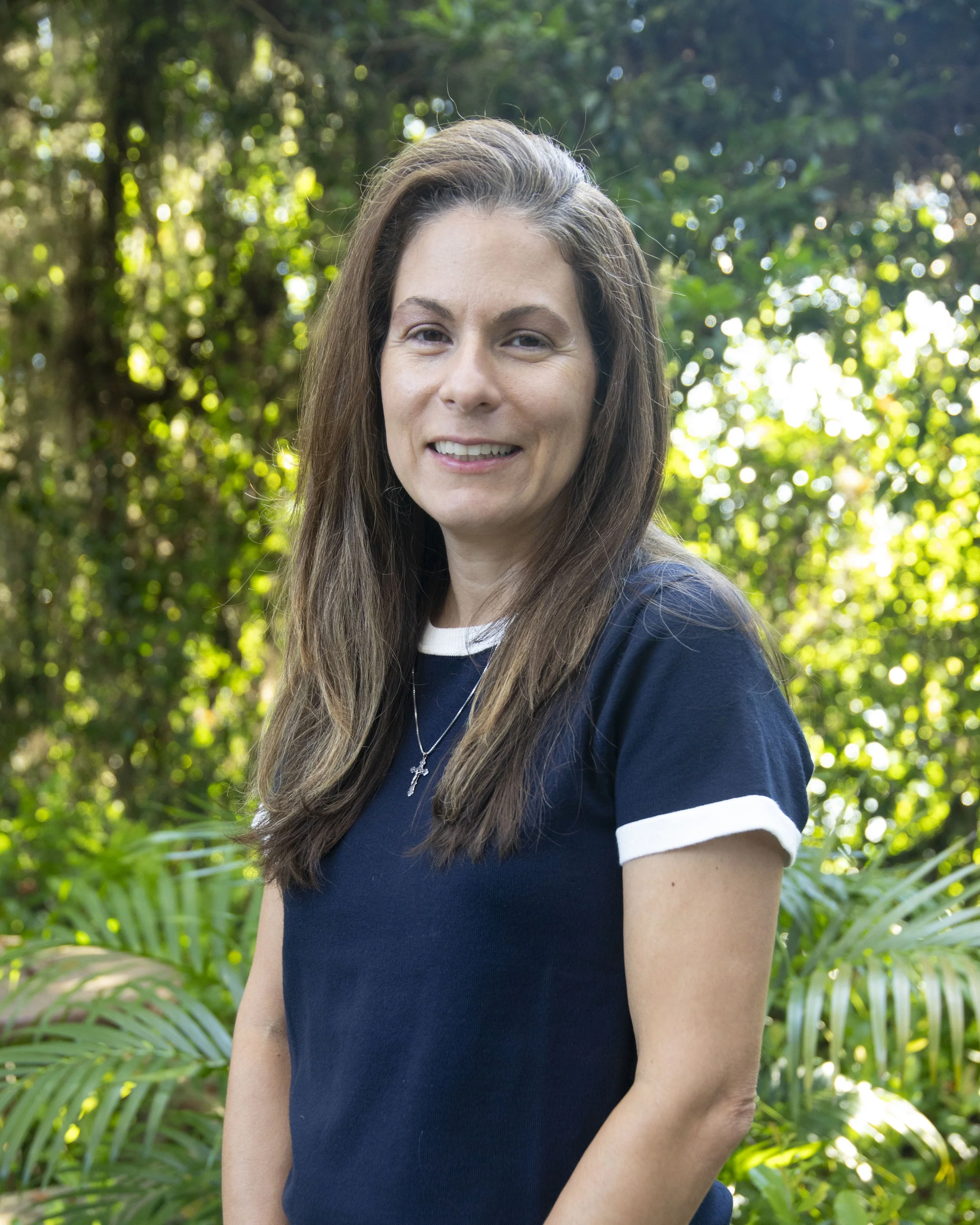 A woman with long brown hair and a cross necklace standing outdoors in front of green foliage, smiling at the camera.