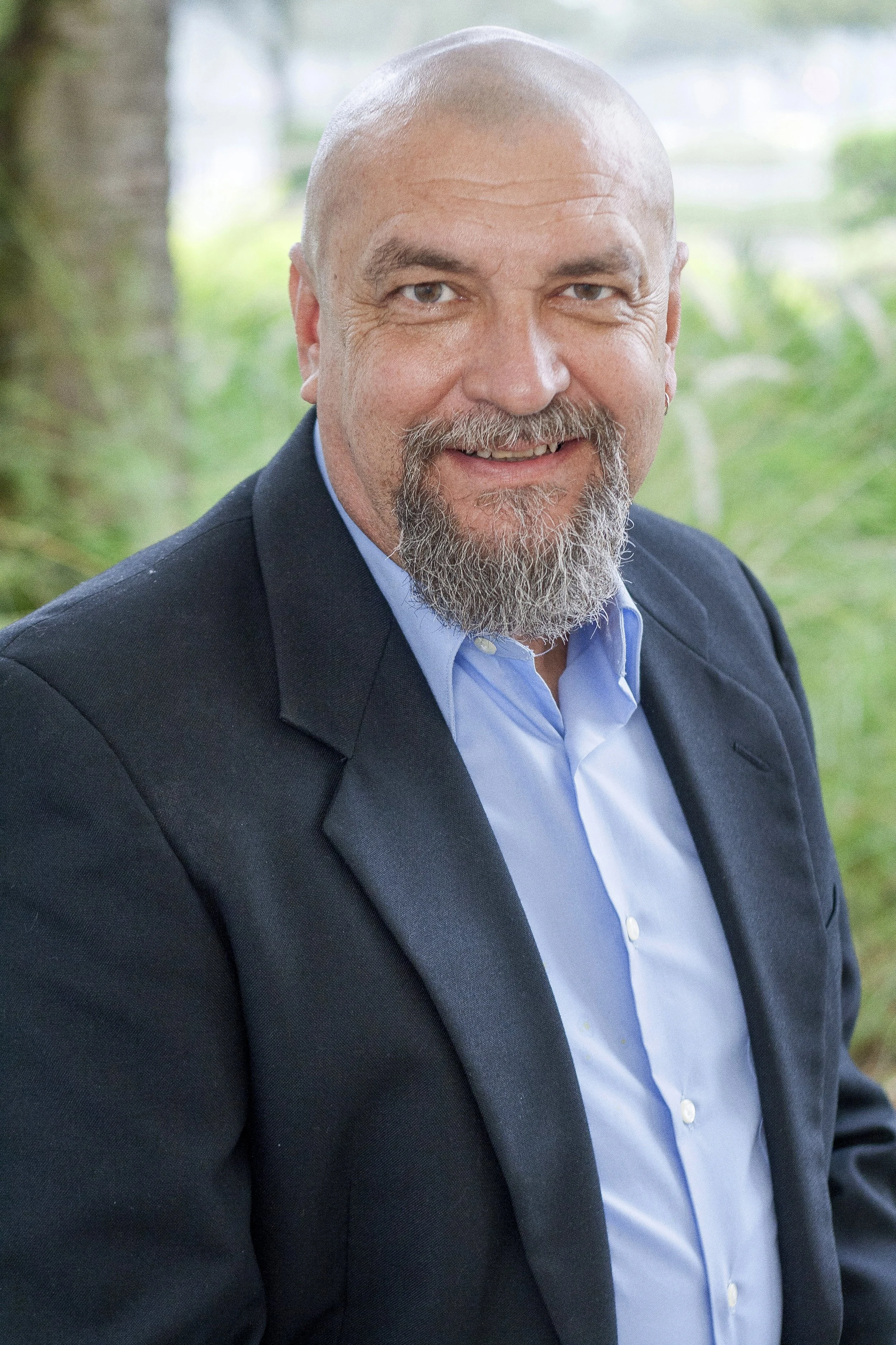 A smiling middle-aged man with a beard and short hair, wearing a dark suit and light blue shirt, sitting outdoors with a blurred green background.