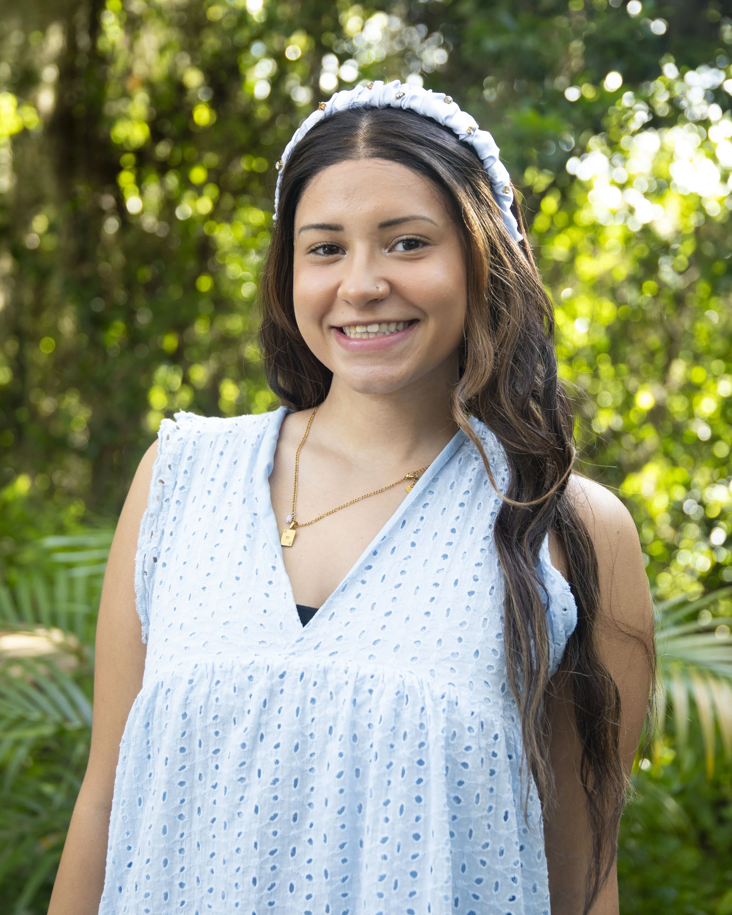 A young woman smiling outdoors in a garden with green foliage, wearing a light blue sleeveless top, a white headband with small decorative beads, and a gold necklace with a small pendant.