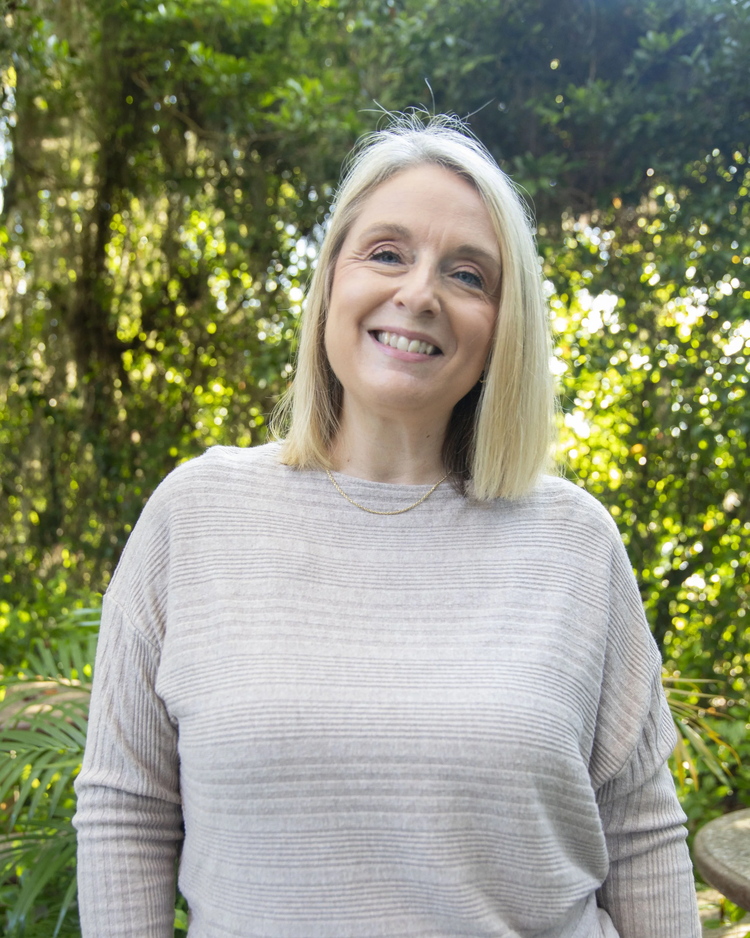 A smiling woman with blonde hair standing outdoors in front of green trees and plants during daytime.