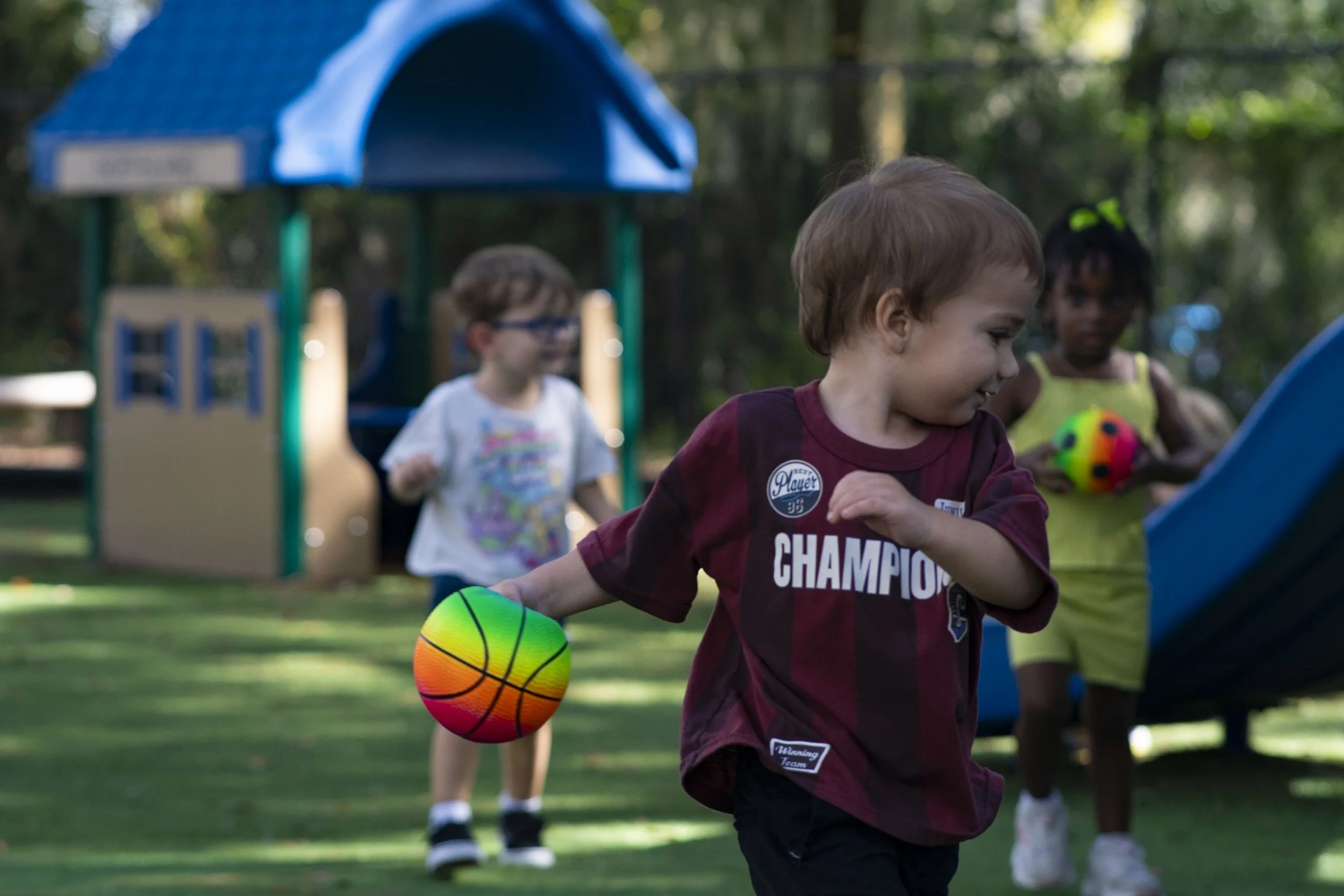 The image shows young children playing with a colorful ball outdoors at a playground surrounded by trees.