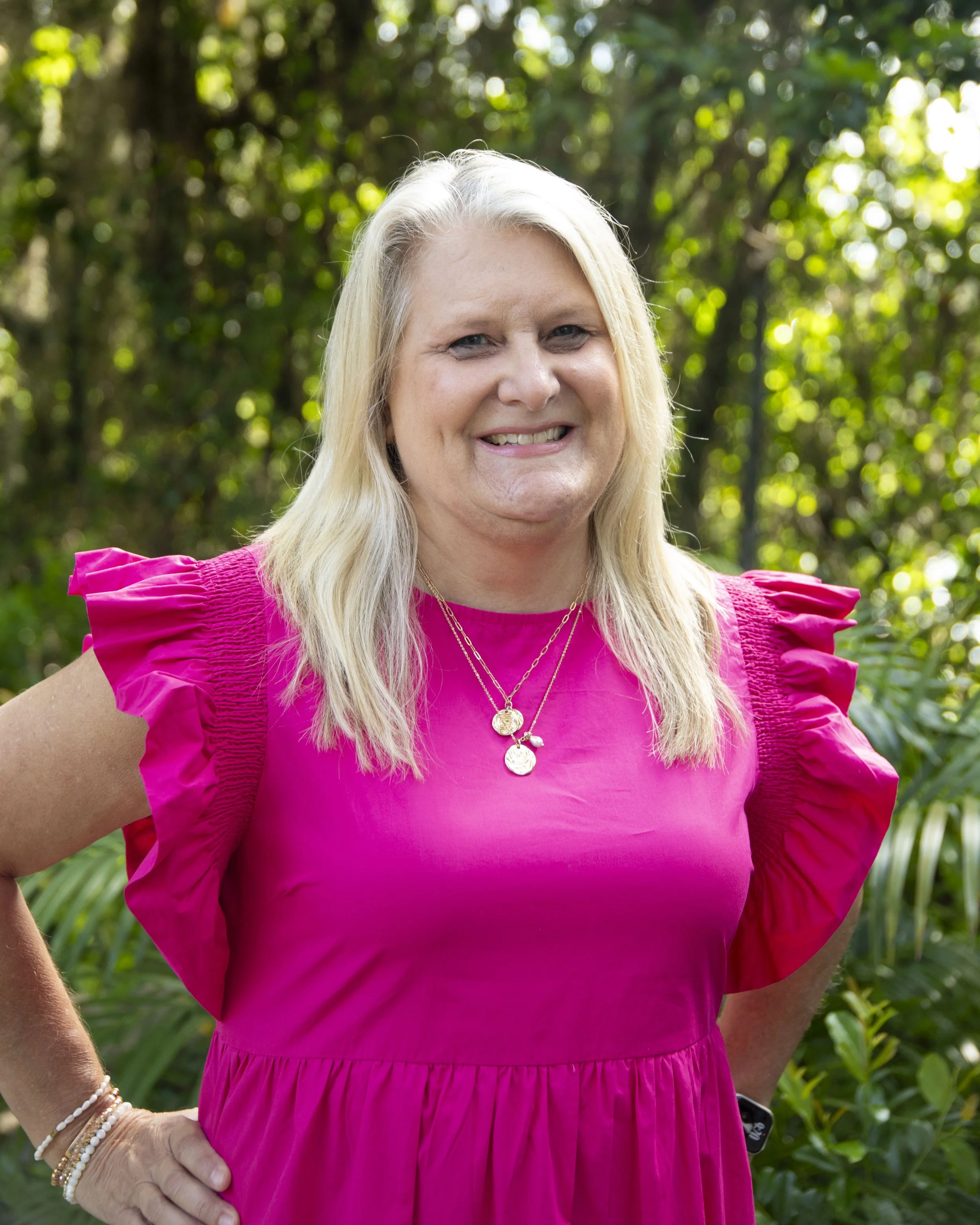 A woman in a vibrant pink dress with puffed short sleeves stands outdoors in a lush, green environment. She has long, blonde hair and is smiling, wearing layered gold necklaces with circular pendants and multiple bracelets on her left wrist.