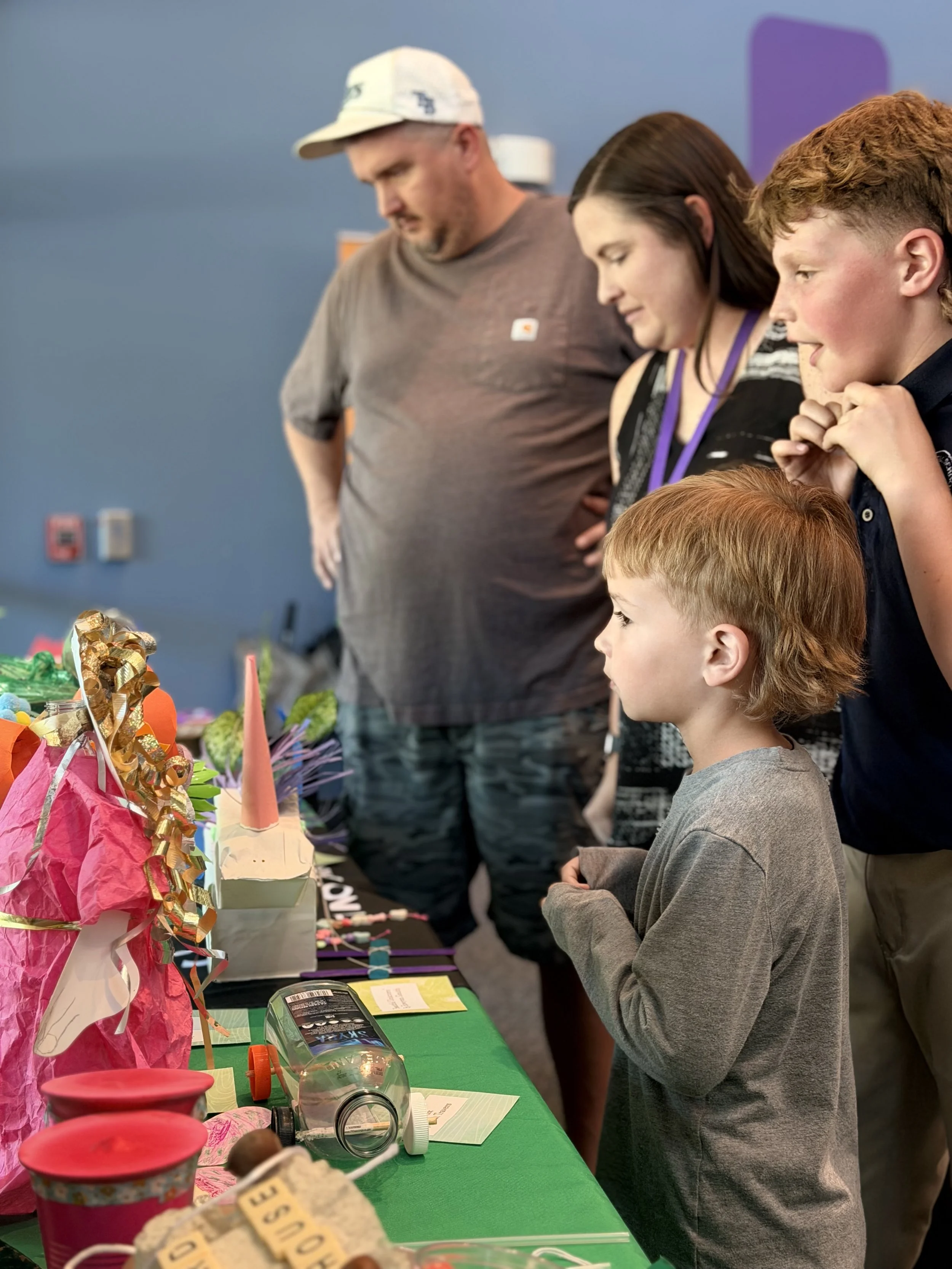 People at a display table, observing colorful items including tissue paper crafts, a small figurine, and a glass dome, with a person using a smartphone nearby.