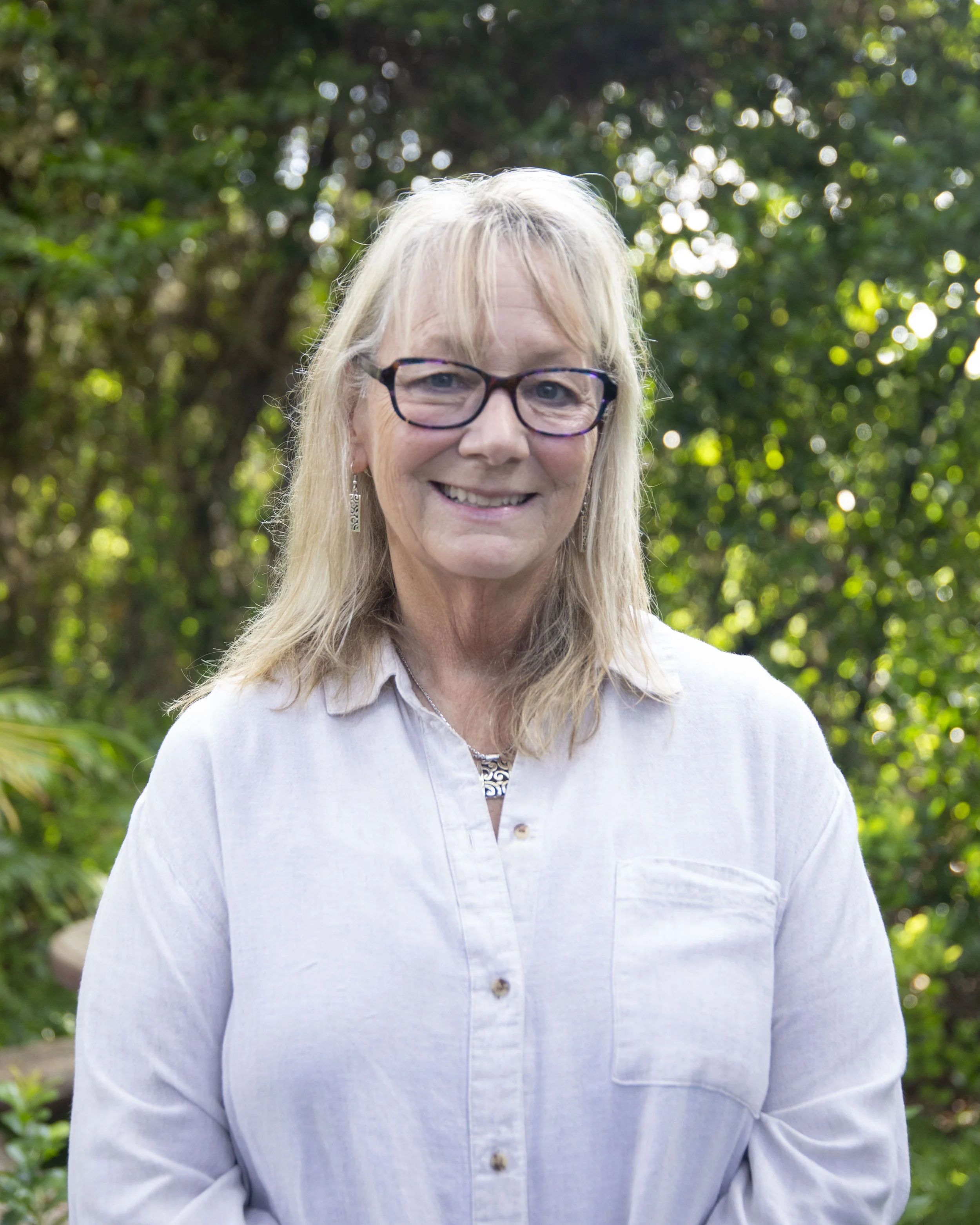 A middle-aged woman with long blonde hair, wearing glasses and a white button-up shirt, smiling outdoors with a blurred green foliage background.