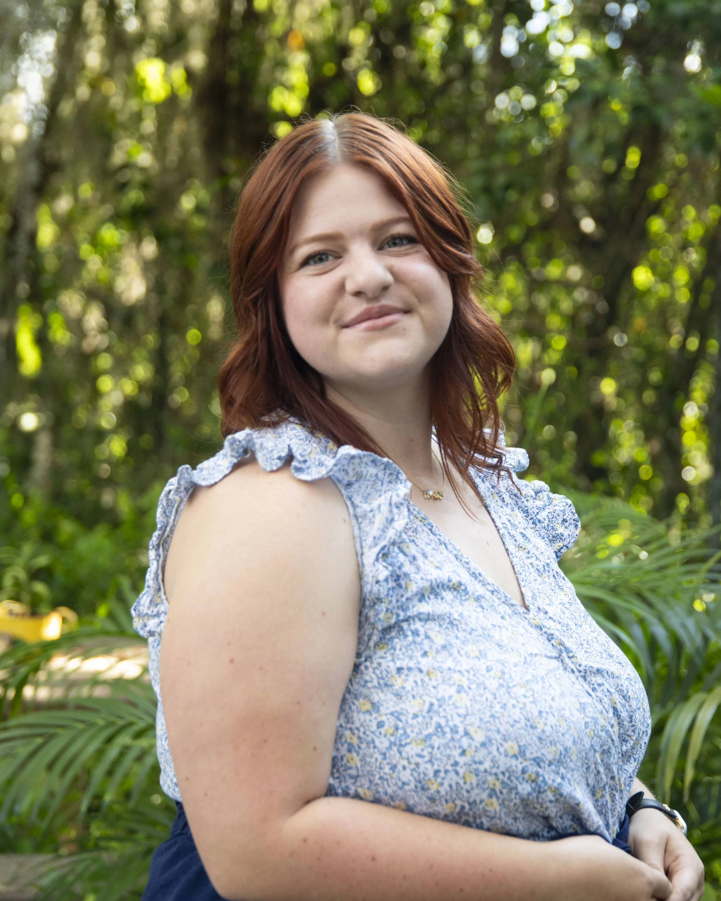 A young woman with reddish-brown hair smiling softly in an outdoor setting with greenery in the background, wearing a blue floral top and a delicate necklace.