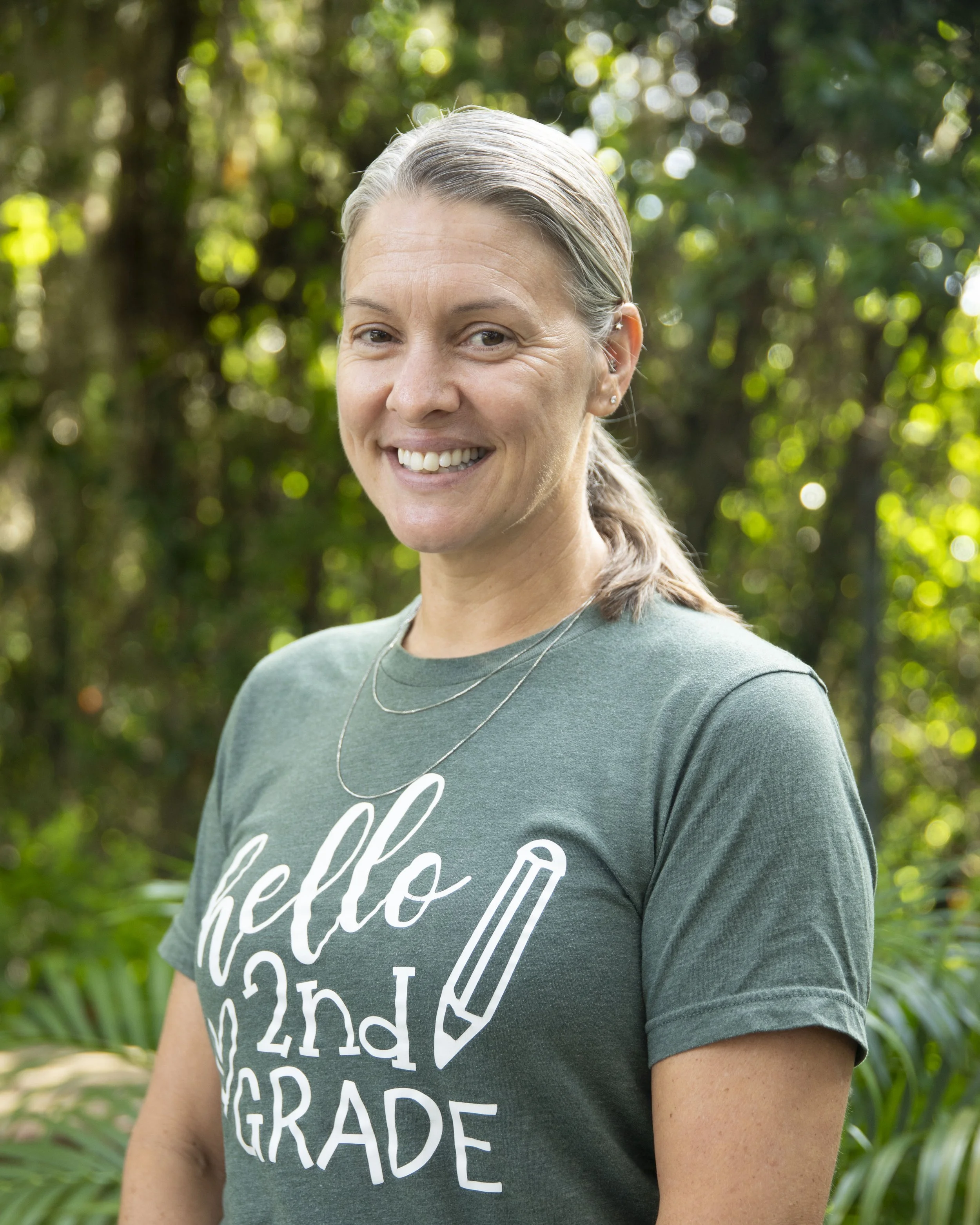 A woman with gray hair and earrings smiling outdoors, wearing a green t-shirt with white text, in a green, leafy environment.