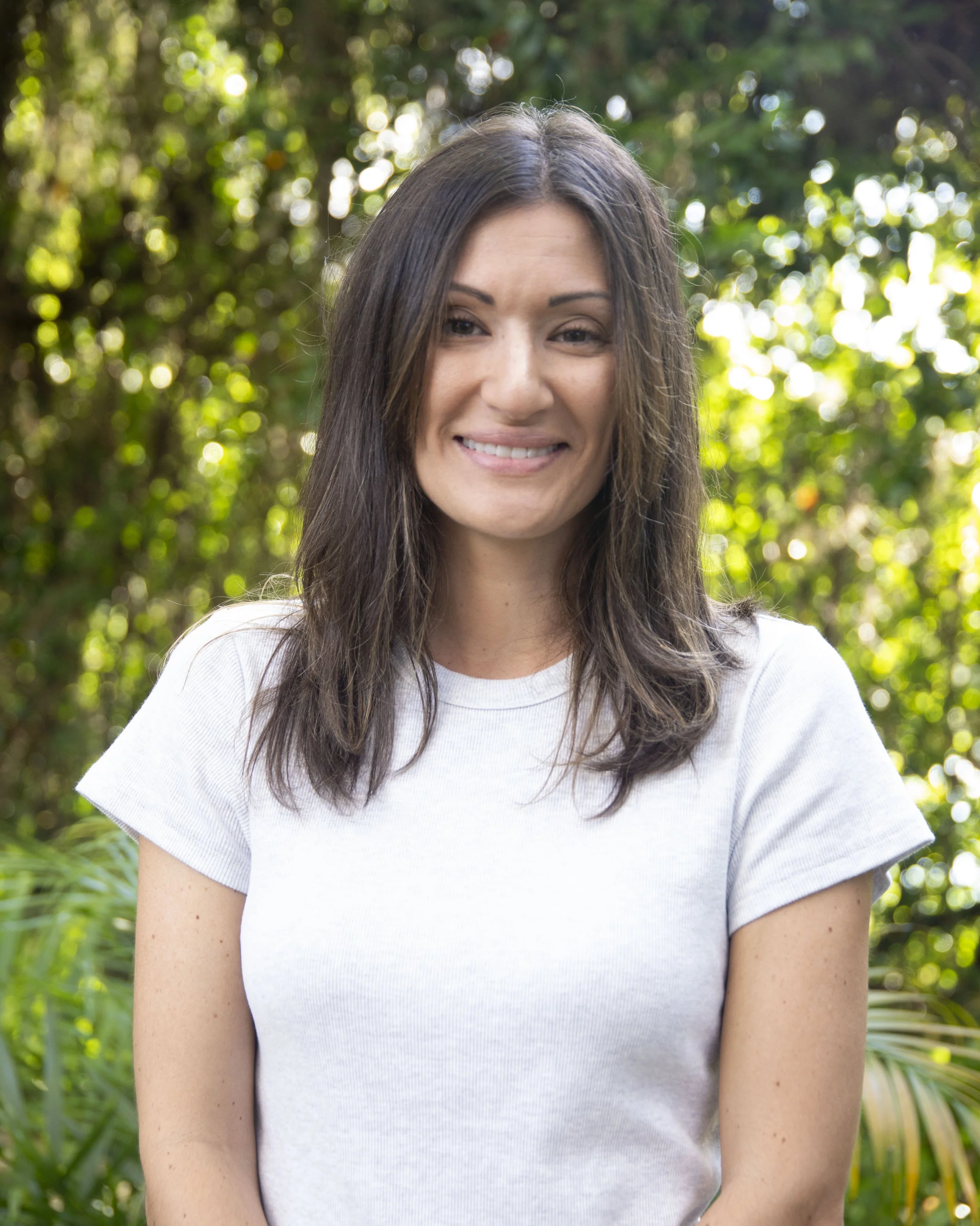 A woman with long brown hair and light skin smiling outdoors in front of green foliage, wearing a white short-sleeved shirt.