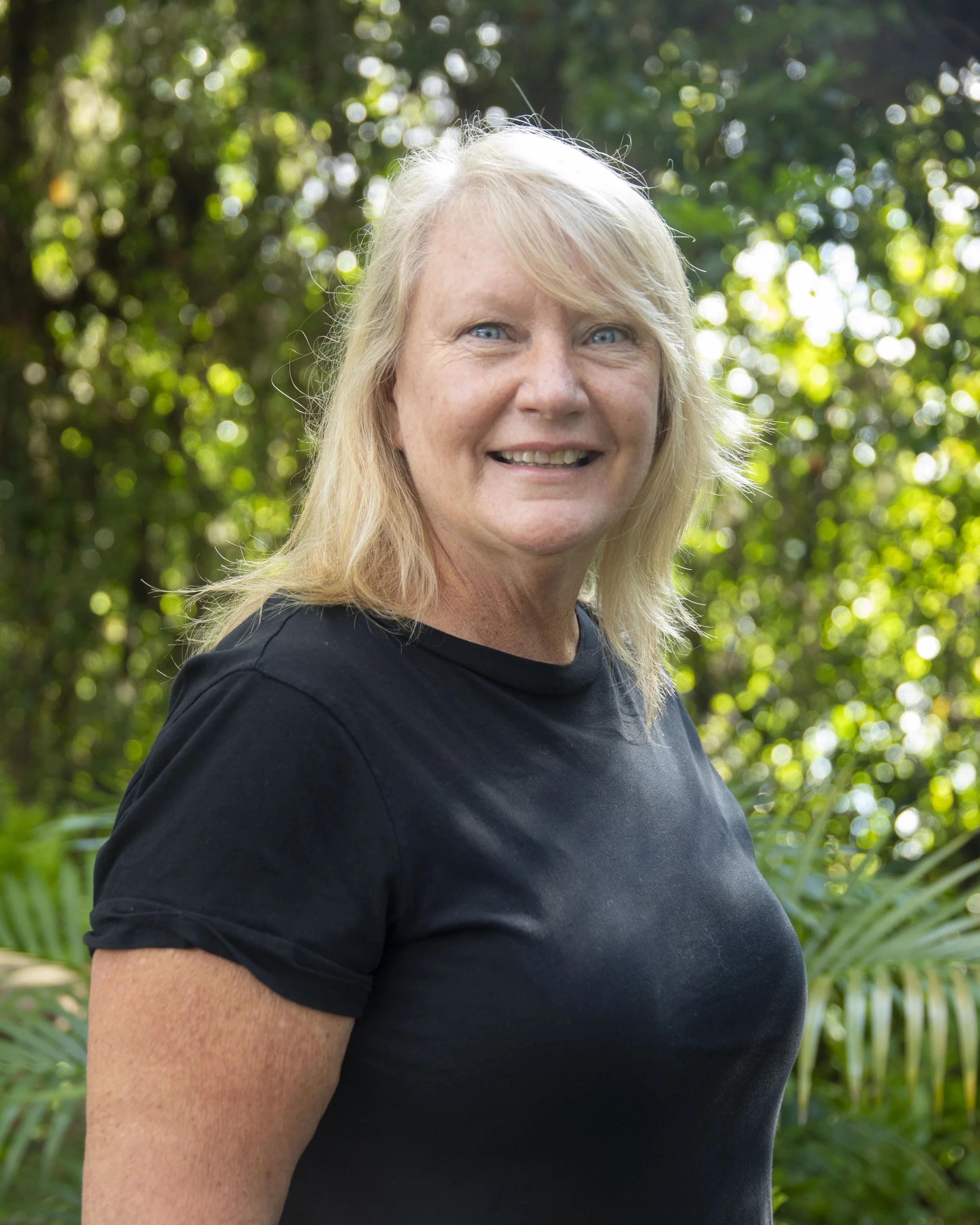 A woman with long blonde hair, blue eyes, and a smile, wearing a black shirt, standing outdoors with lush greenery in the background.