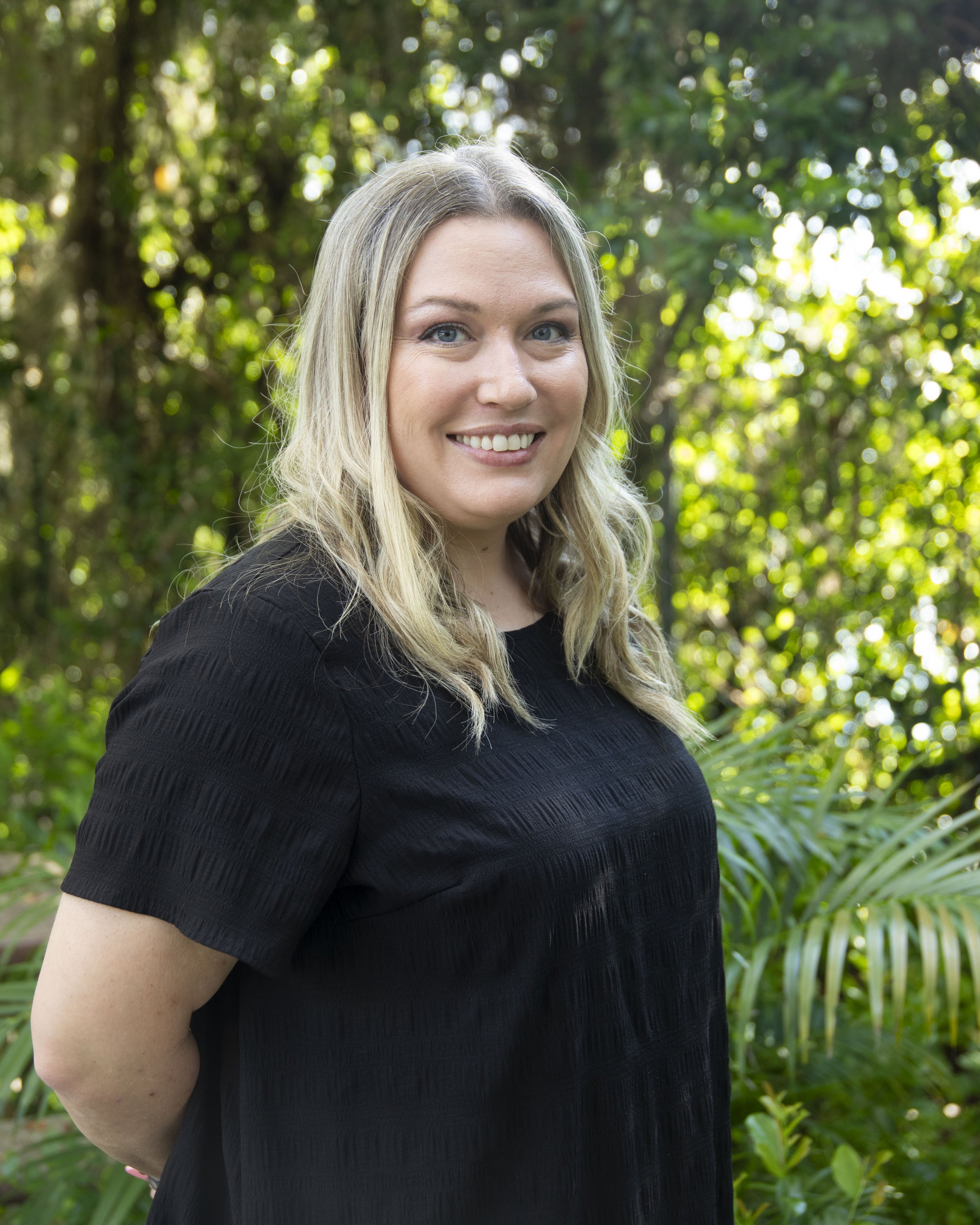 A woman with long blonde hair and blue eyes smiling outdoors, wearing a black top, with green foliage and trees in the background.