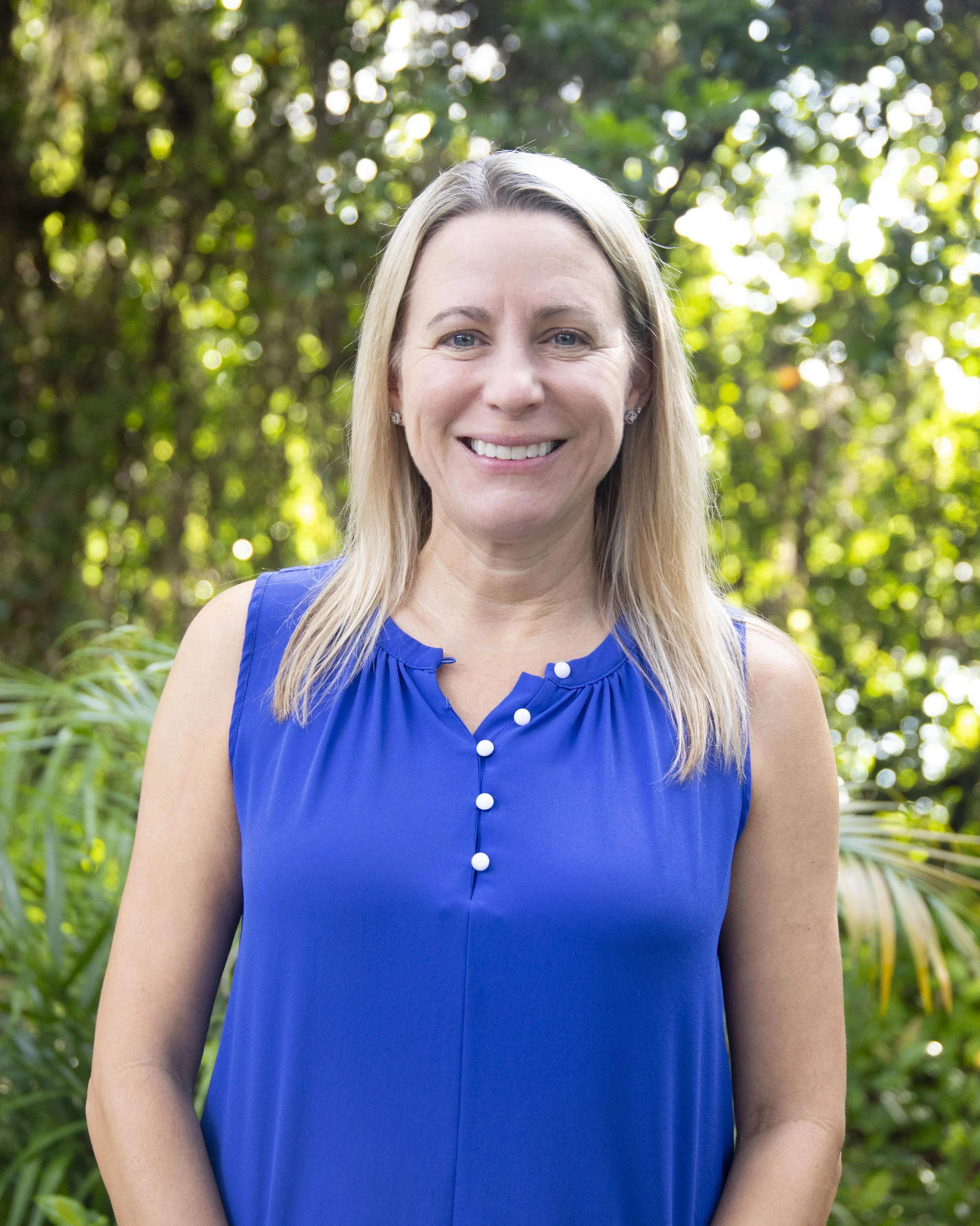A woman with long blonde hair, blue eyes, and a bright smile, wearing a sleeveless bright blue dress with white buttons, standing outdoors in front of a background of green trees and foliage.