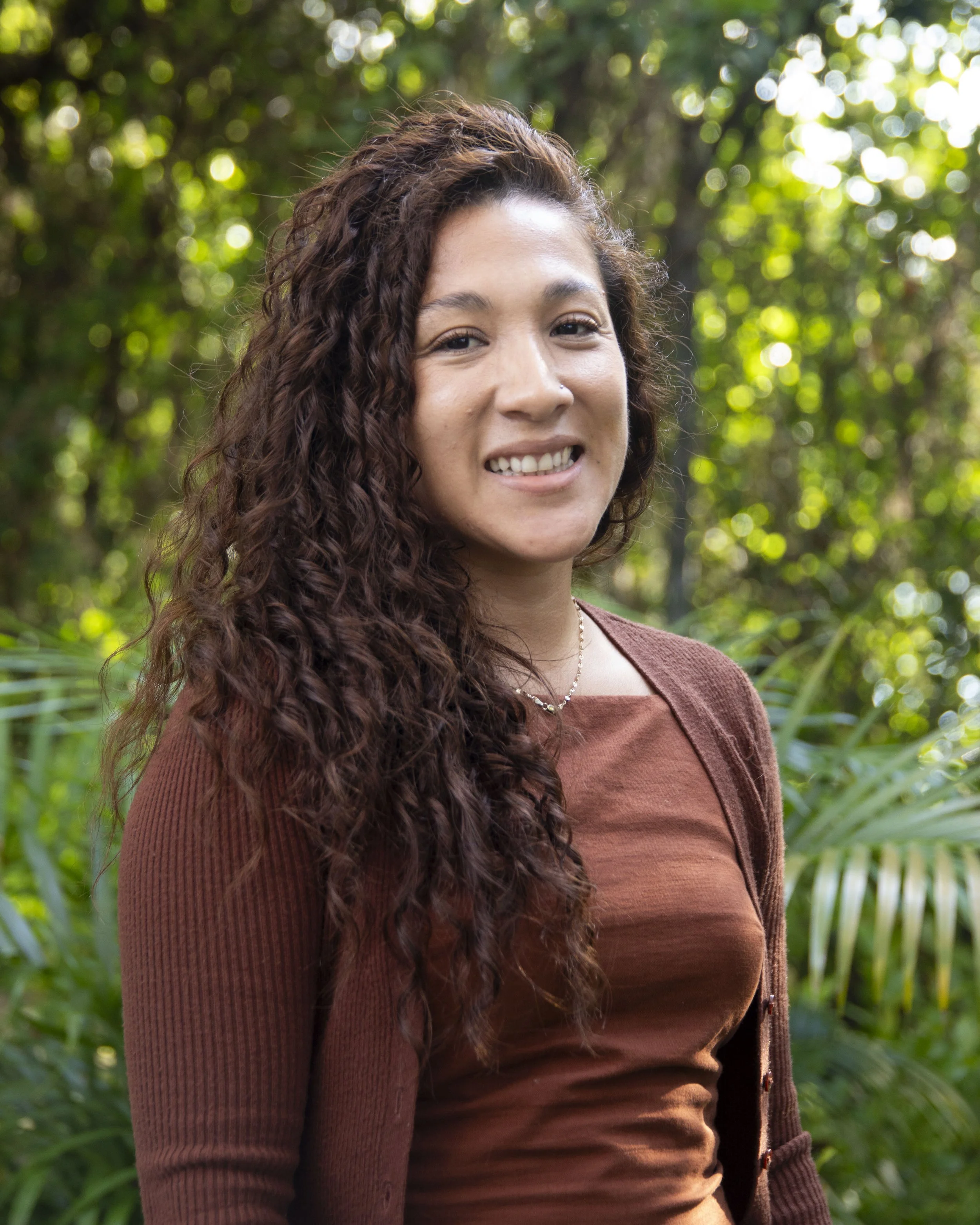 A woman with long, curly brown hair smiling outdoors in a lush, green environment.
