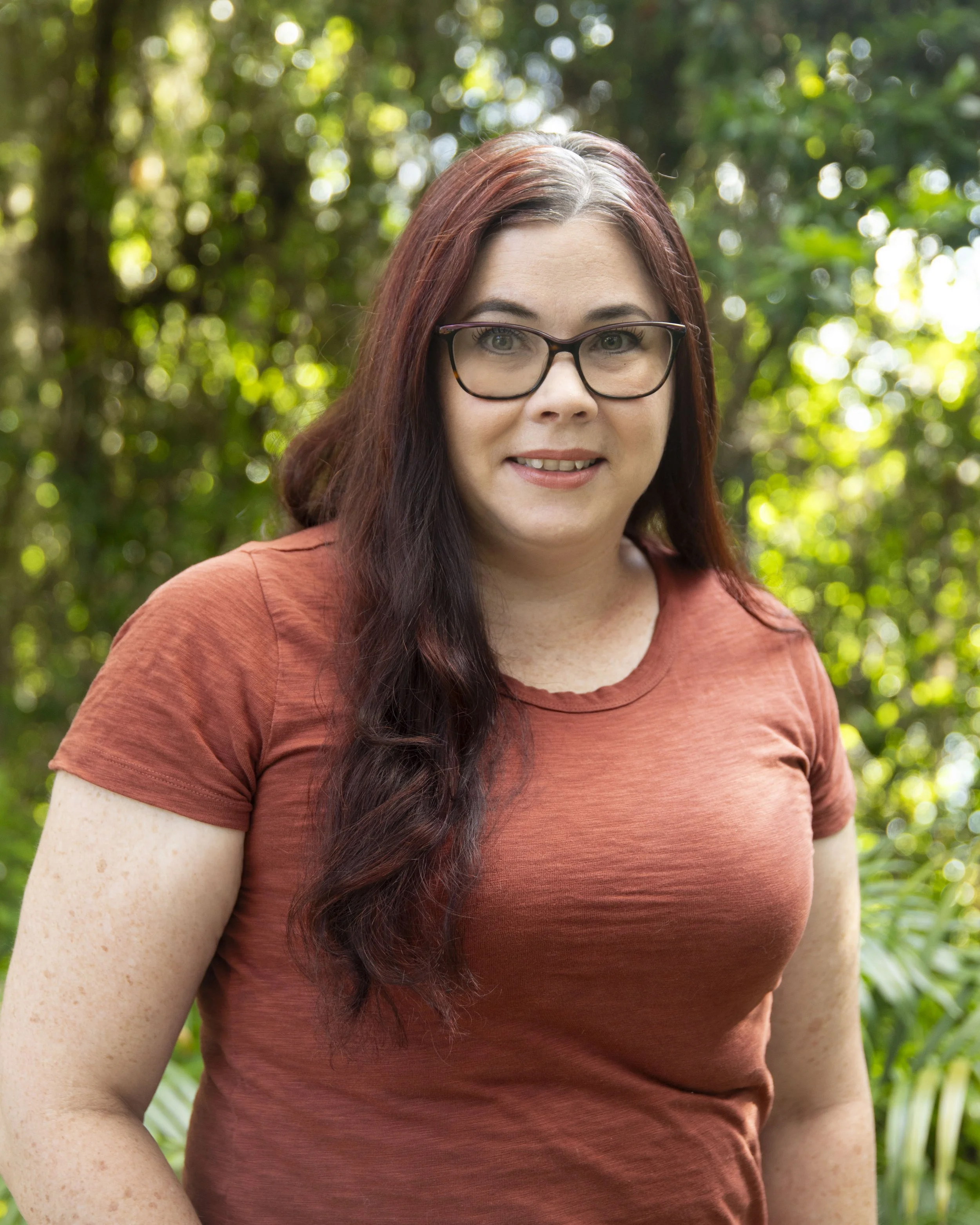 A woman with long dark red hair, wearing glasses, shows her smiling outdoors with blurred green foliage in the background.