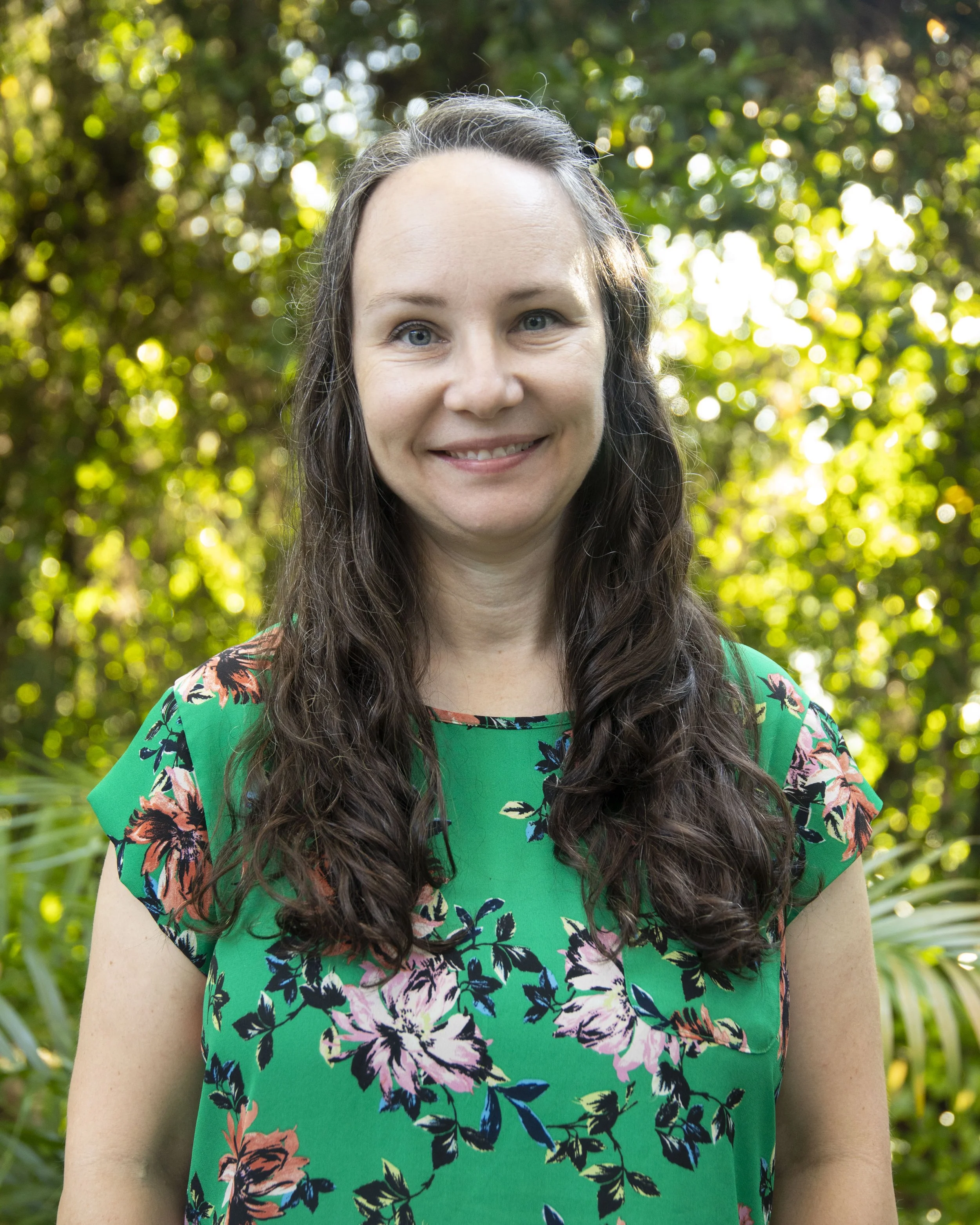 A woman with long wavy brown hair and blue eyes standing outdoors against a blurred background of green trees and sunlight. She is wearing a green dress with a floral pattern.