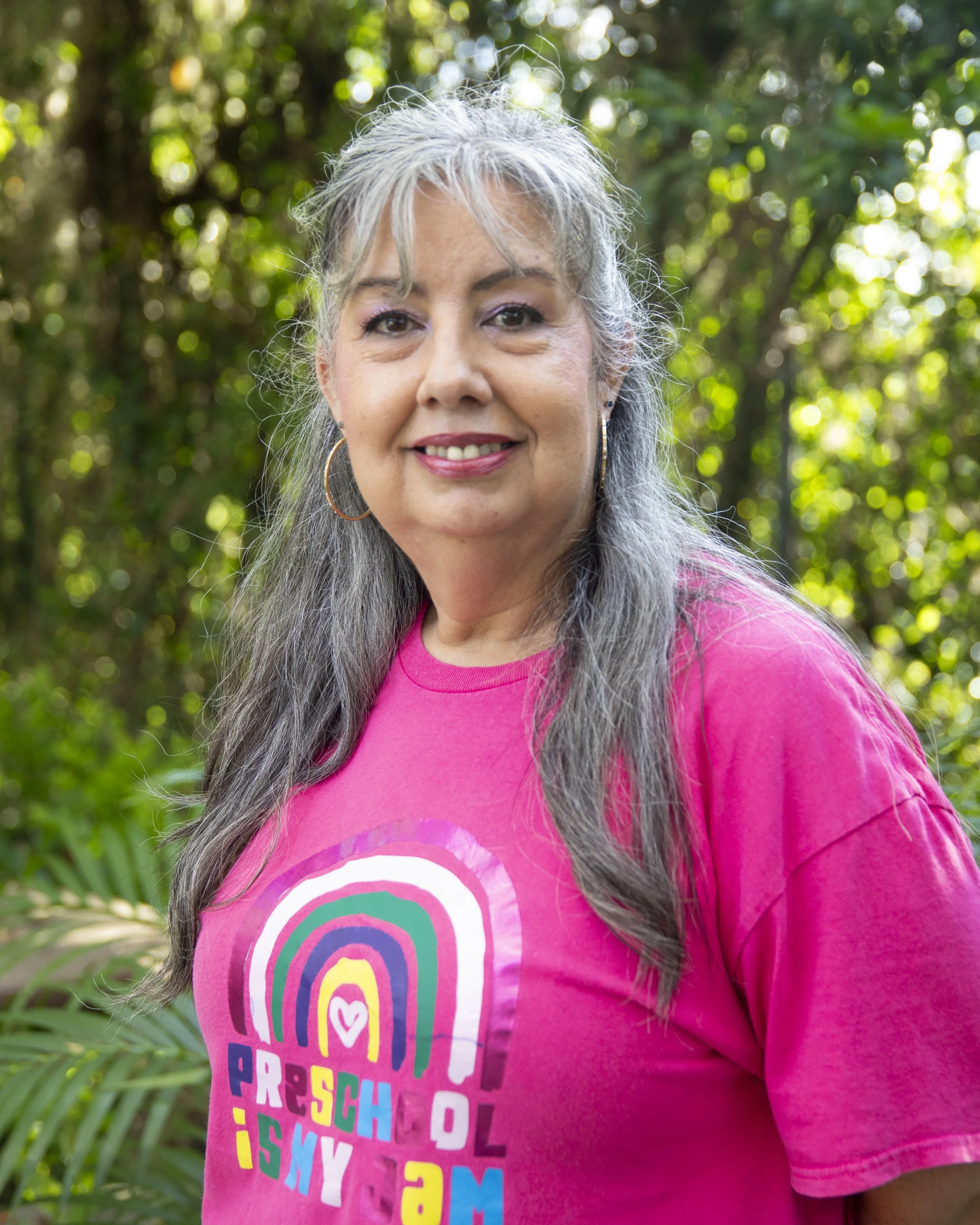 A woman with gray hair and earrings is smiling outdoors, wearing a pink t-shirt with colorful rainbow text, against a background of green foliage.