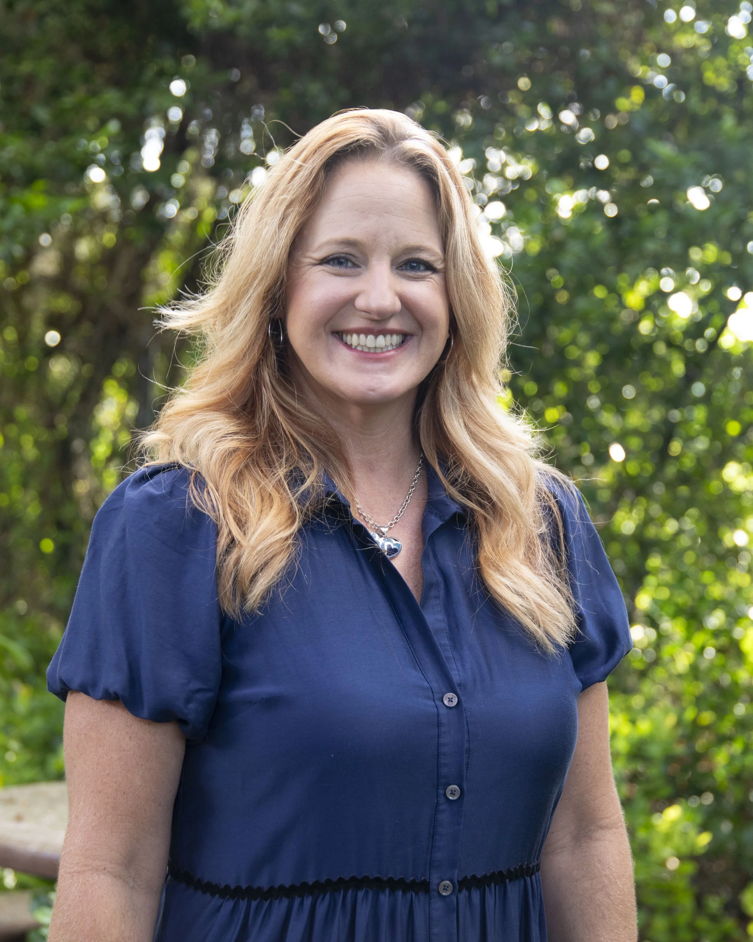 A woman with long reddish-blonde hair, wearing a blue dress with buttons and a necklace, smiling outdoors with a background of green trees and sunlight.