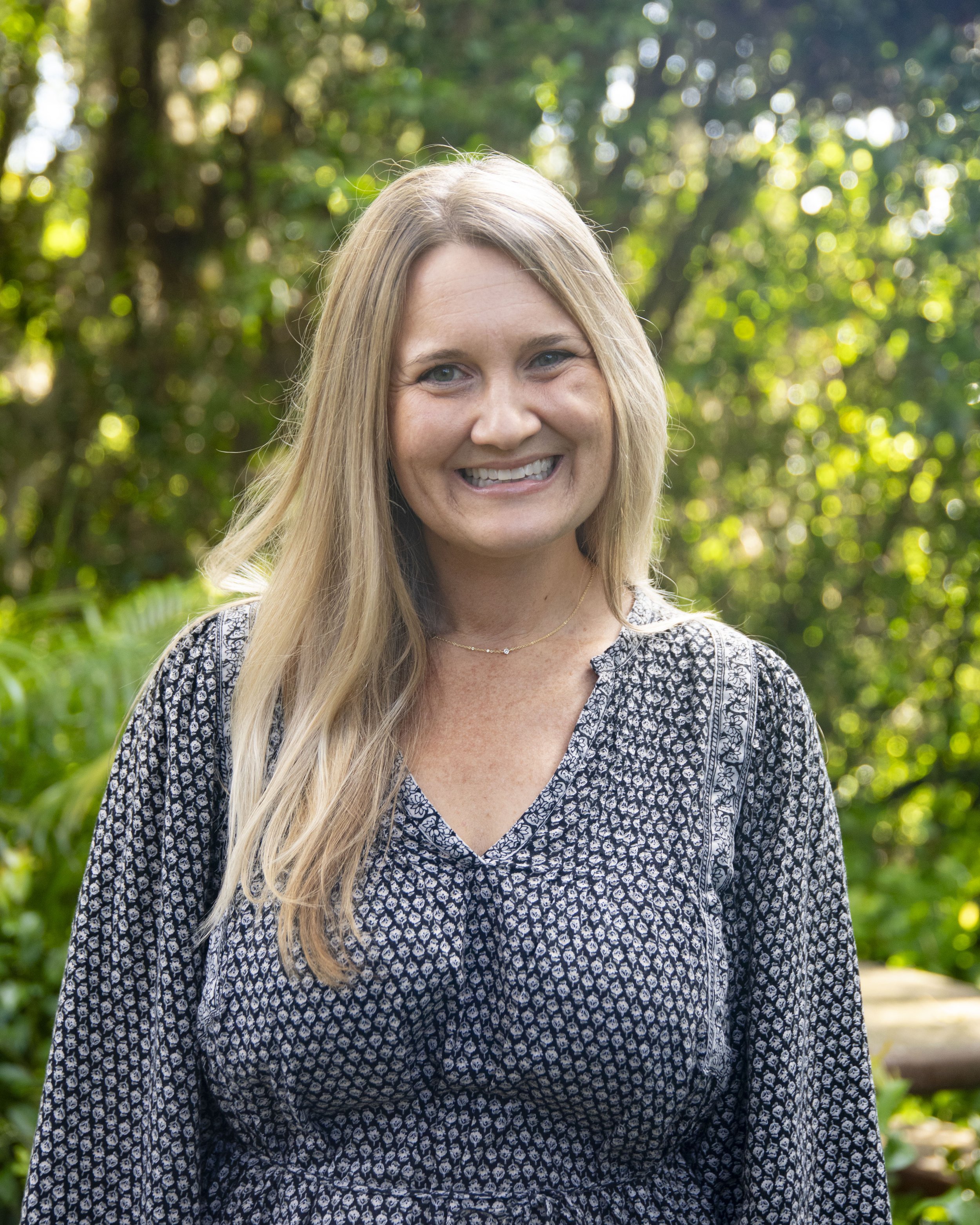 A woman with long blonde hair smiling outdoors with a background of green trees and sunlight.