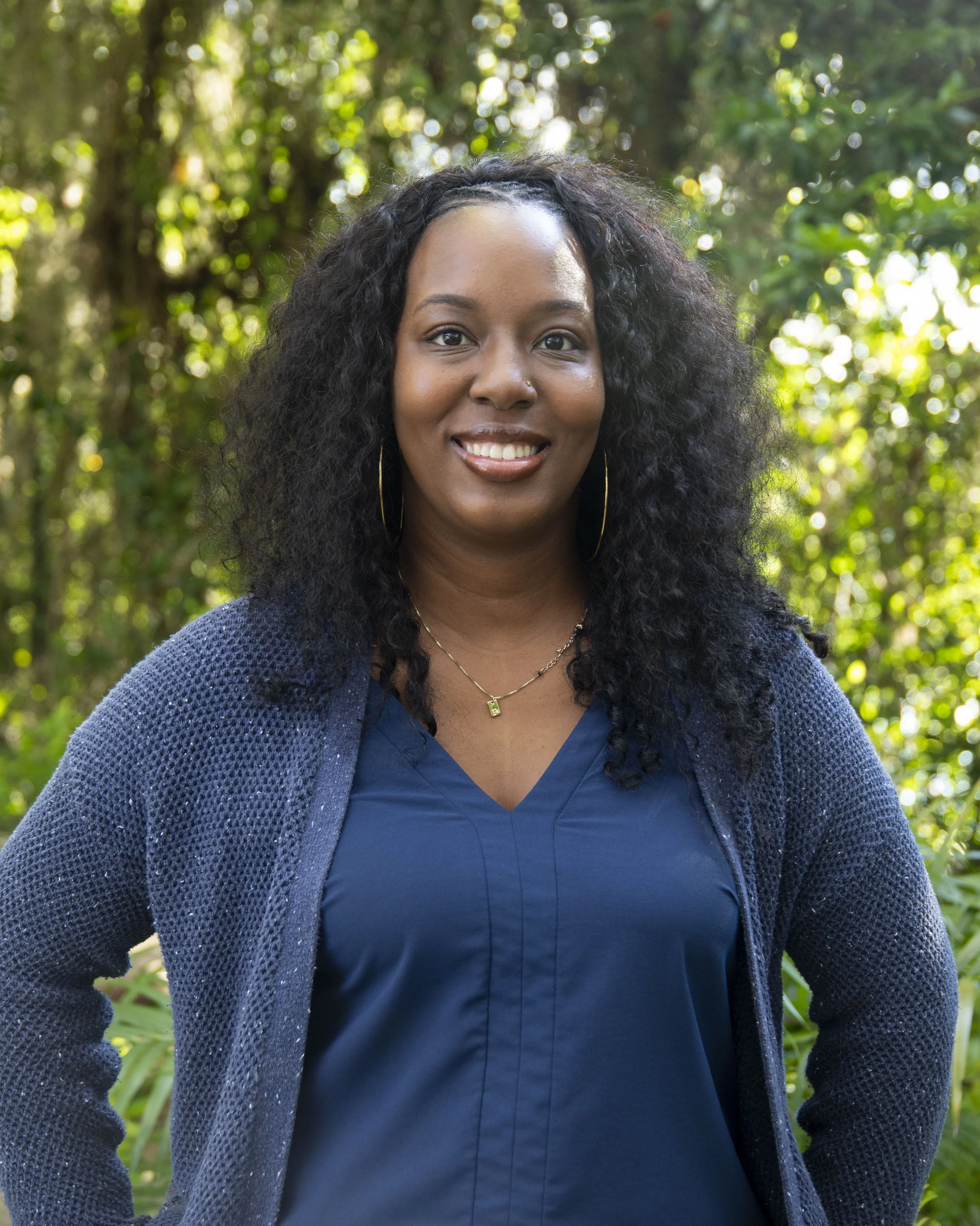 Portrait of a smiling African American woman with curly black hair, wearing a blue shirt, a dark blue knitted cardigan, a gold necklace, and hoop earrings, standing outdoors with blurred green foliage background.