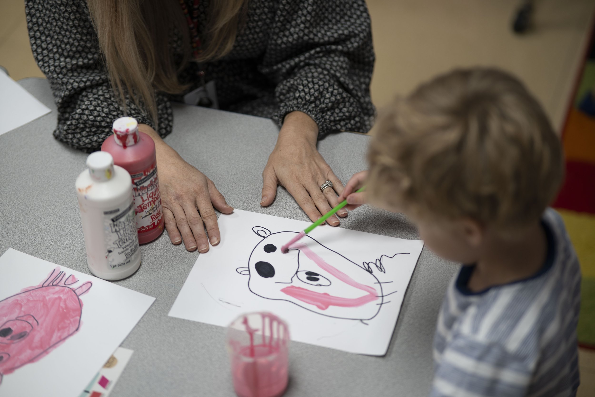 A young child and an adult sitting at a table engaged in a craft activity, coloring and drawing pictures on paper, with bottles of paint or glue on the table.
