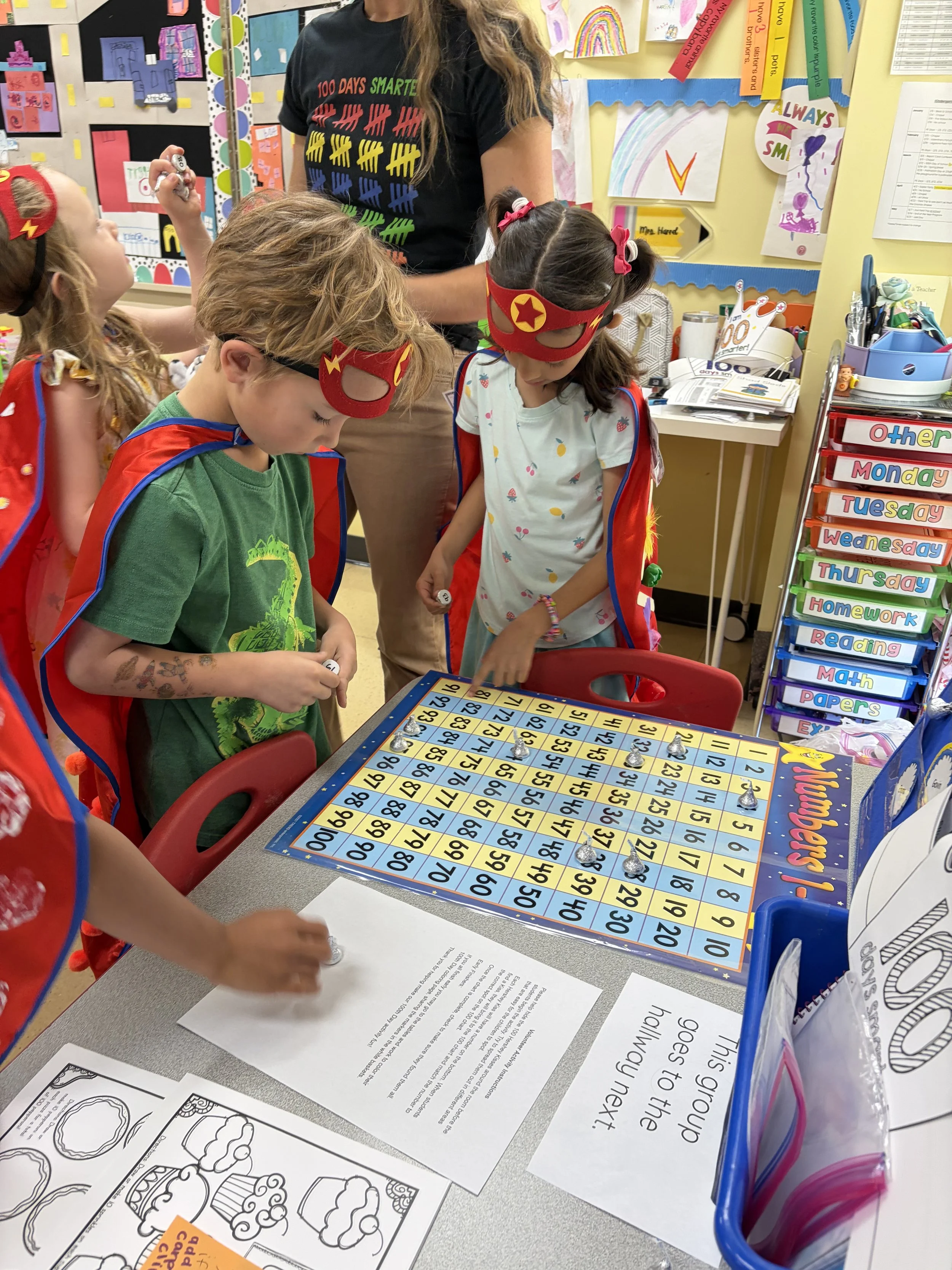 Colorful classroom with children wearing superhero masks around a table, playing a number game on a board, with worksheets and art projects displayed on the walls.