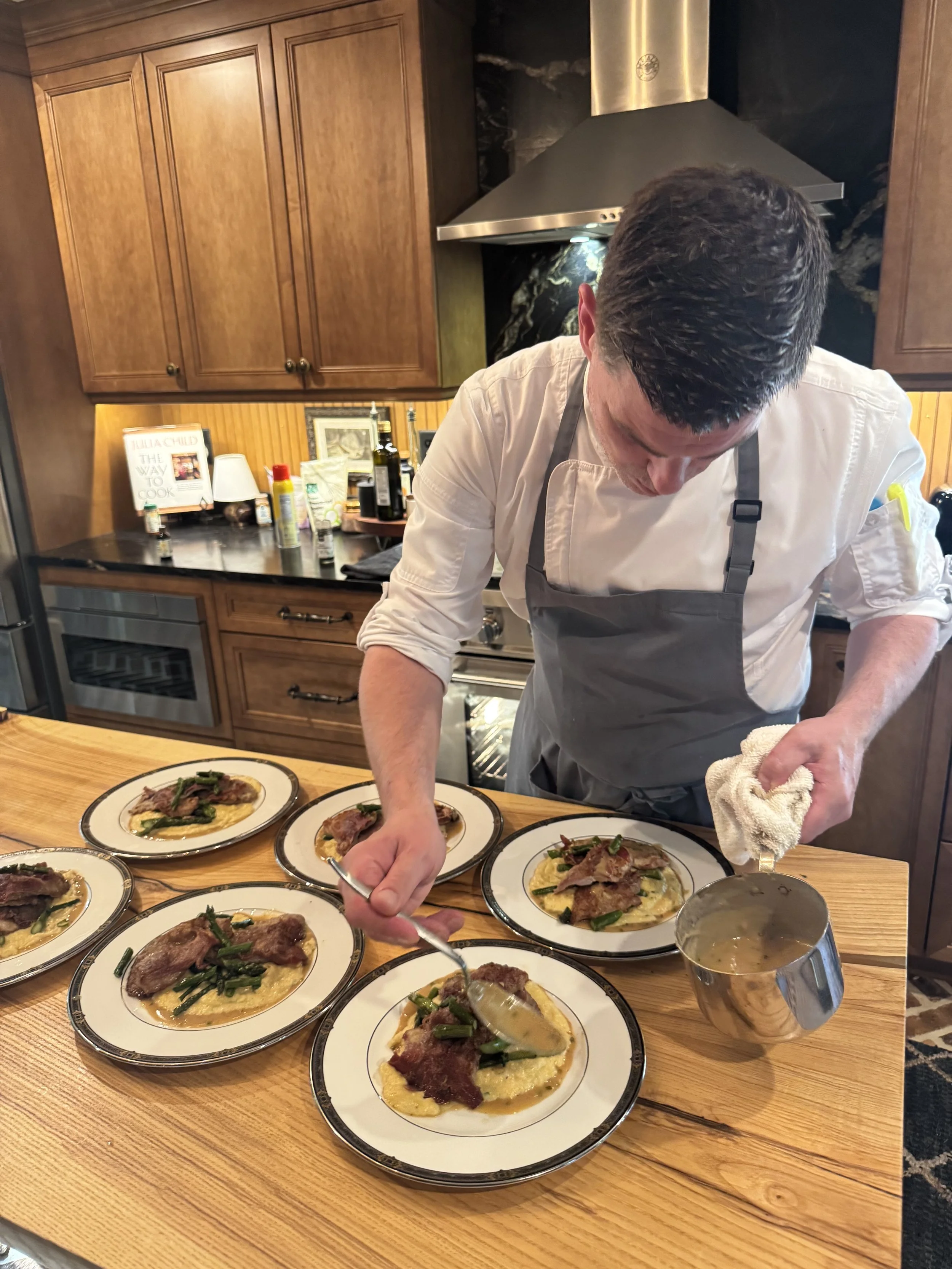 A man wearing a white shirt and gray apron is garnishing multiple plates of a dish with a creamy yellow sauce, served with vegetables and slices of meat, in a wooden kitchen. There are several plates arranged on a wooden table, and the man is holding a spoon and a towel.