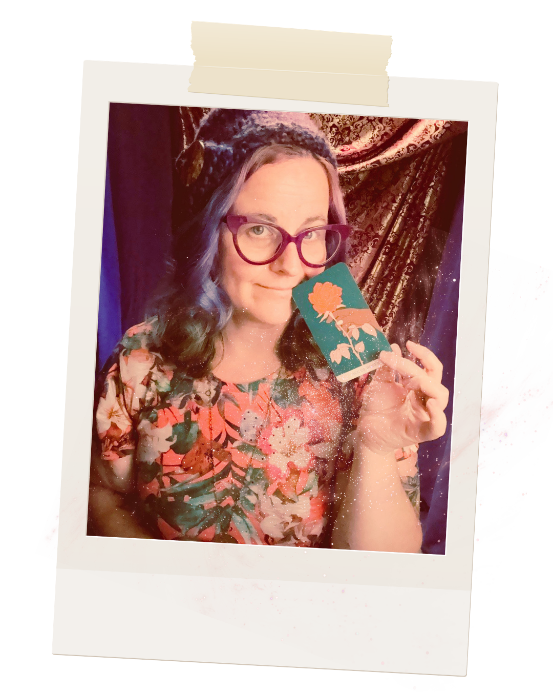 A woman with purple hair, glasses, and a floral top holding a card with a flower illustration, posing in front of a patterned curtain.