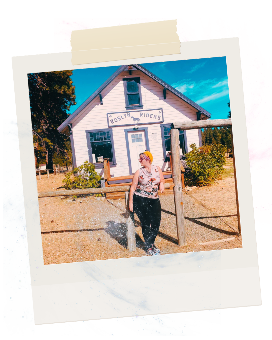 Woman wearing sunglasses, floral sleeveless top, and orange headband standing in front of the Roslyn Riders historic building with a sign, trees, and a bright blue sky.