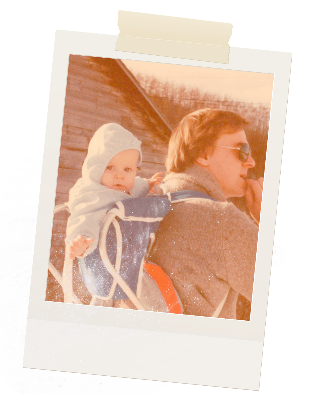 A woman carrying a young child on her back outdoors during sunset, with the child wearing a hooded jacket. The woman has dark hair and is wearing sunglasses and a coat.