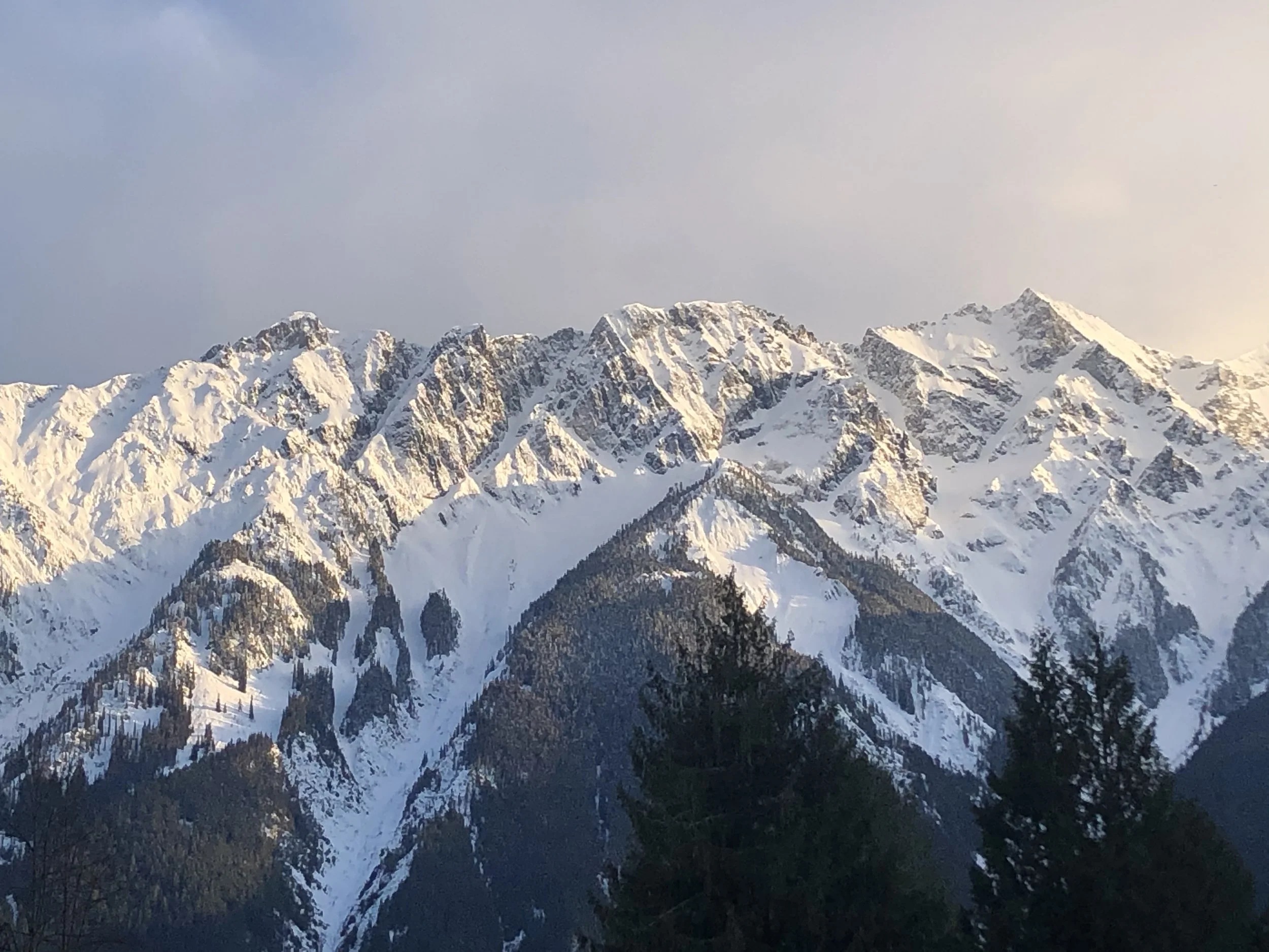 Snow-capped mountain range under a partly cloudy sky with some forested areas at the base.