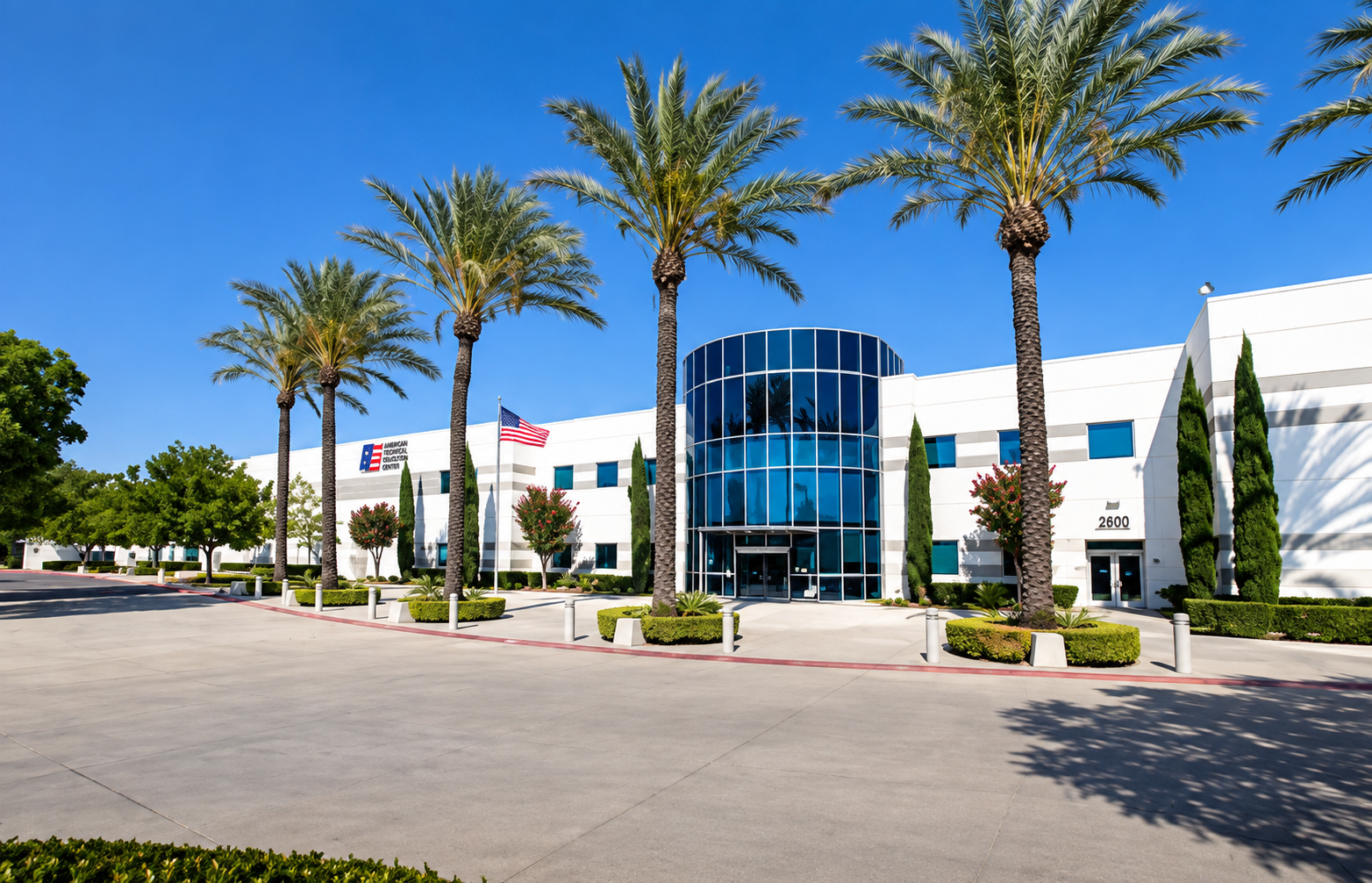 Modern white office building with a curved glass entrance, surrounded by palm trees, smaller trees, and neatly landscaped bushes, under a bright blue sky.