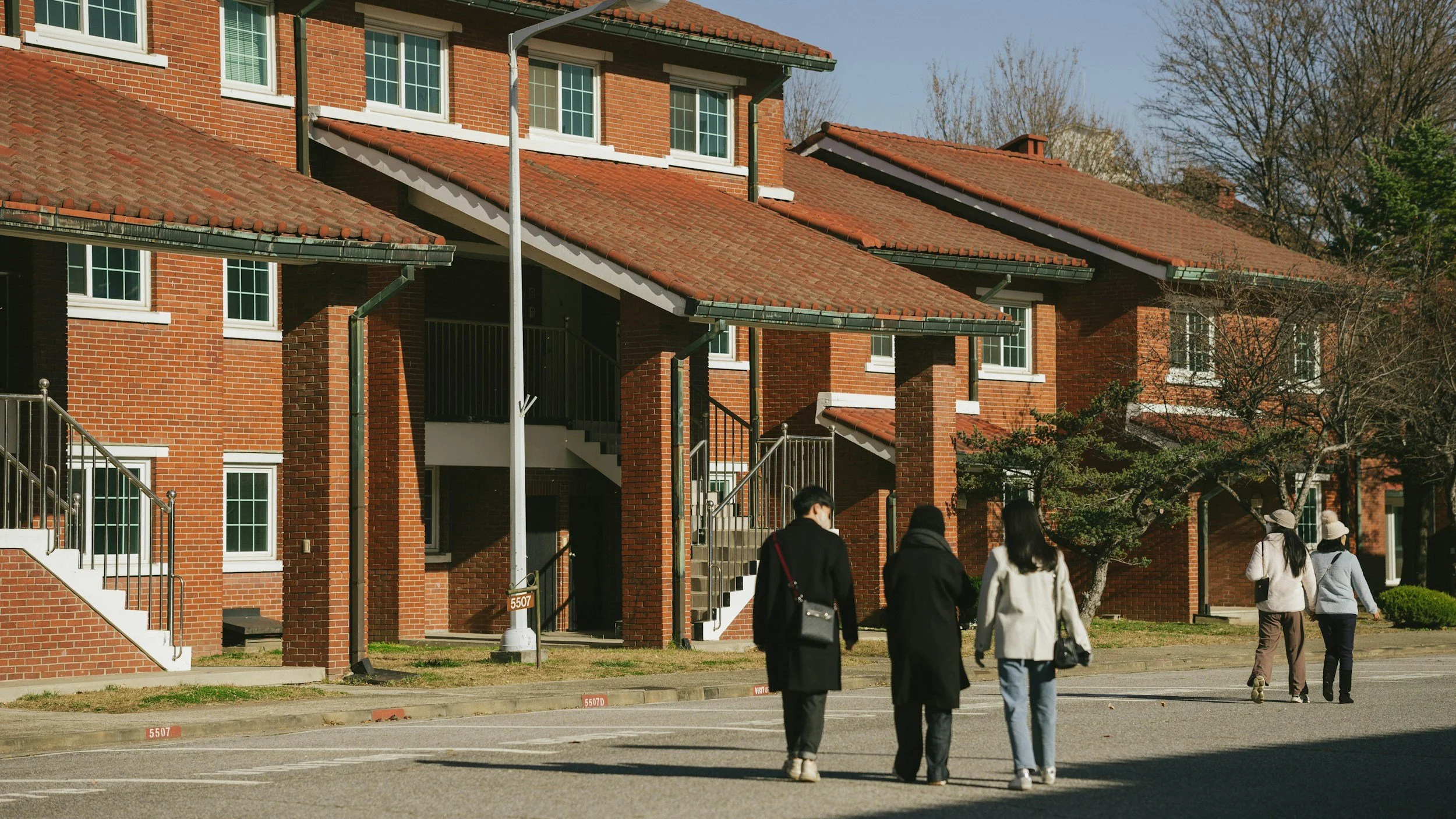 A group of four people walking outside a red brick apartment building on a sunny day. The building has white-framed windows and a staircase leading to the entrance.