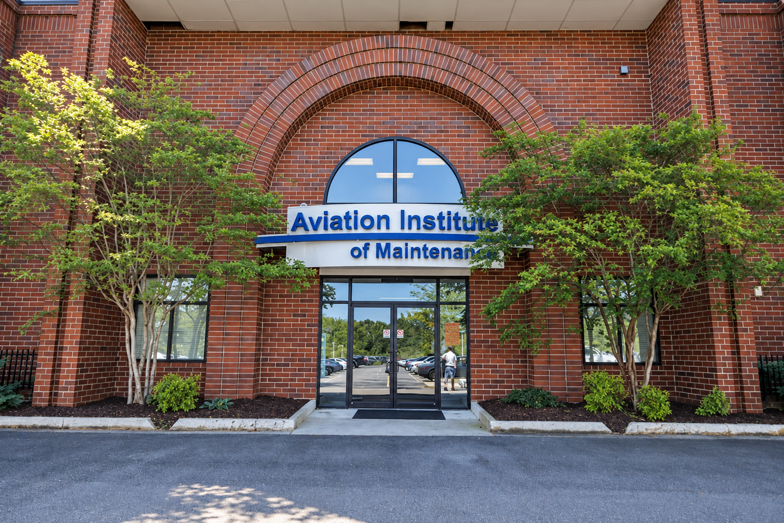 Brick building with sign that reads 'Aviation Institute of Maintenance' above glass doors, surrounded by trees and parking lot.