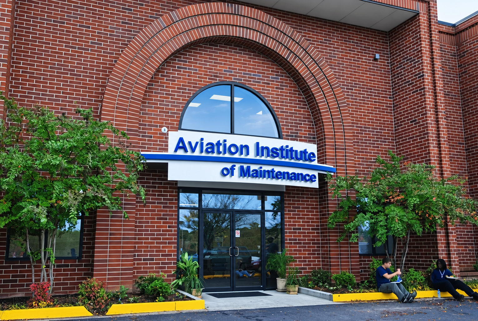 The Aviation Institute of Maintenance building Manassas, Virginia made of red brick, featuring a large window and a sign with blue lettering. Two students sit outside, reading and chatting, with greenery and plants around the entrance.