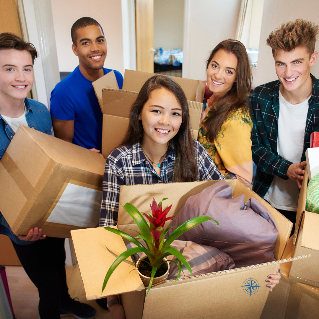 Group of young adults moving into a new home, holding cardboard boxes with belongings, smiling, with a potted plant in a box in the foreground.