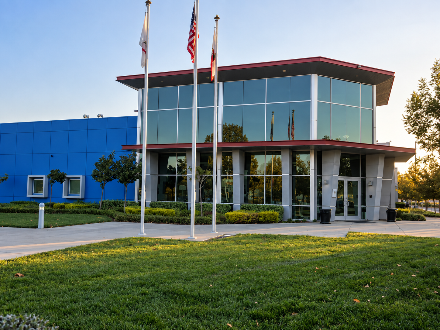 Modern two-story office building with large glass windows, blue and gray exterior, surrounded by green lawn and small trees, with American flags on flagpoles in front, during late afternoon  | Compass Rose Student Housing