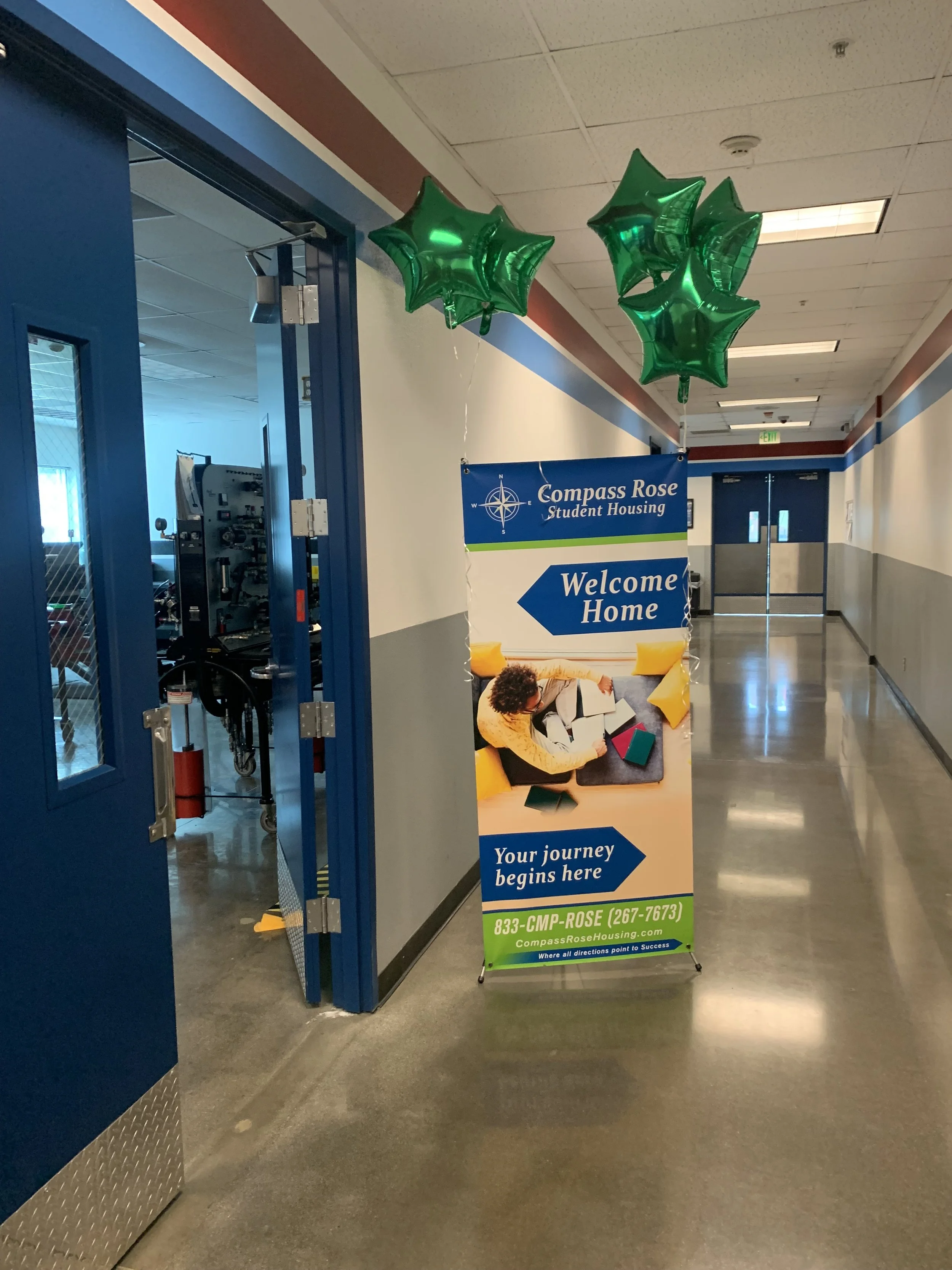 Indoor hallway with a blue door on the left and a sign with green star-shaped balloons attached, indicating the entrance to Compass Rose Student Housing with a welcome message and contact information.
