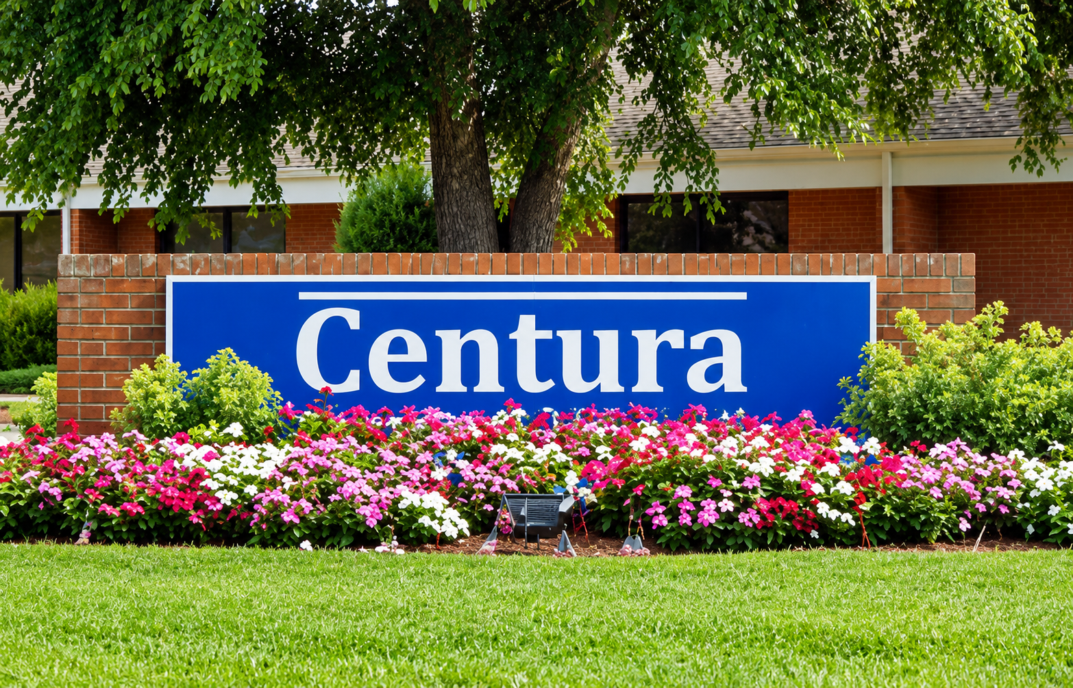 Blue sign with white lettering that says 'Centura' in front of a brick wall, surrounded by pink, white, and purple flowers and green bushes, with a tree and building in the background.