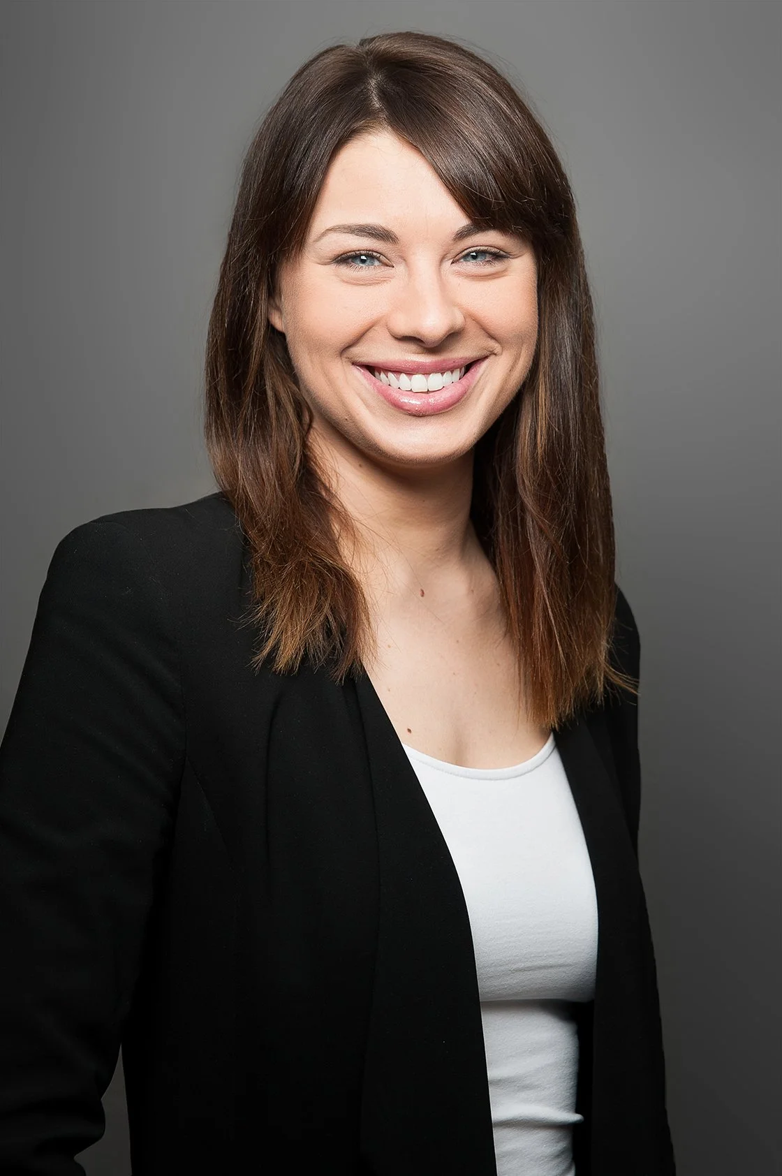 Porträt einer lächelnden Frau mit braunen Haaren, schwarzer Jacke und weißem Oberteil vor grauem Hintergrund.