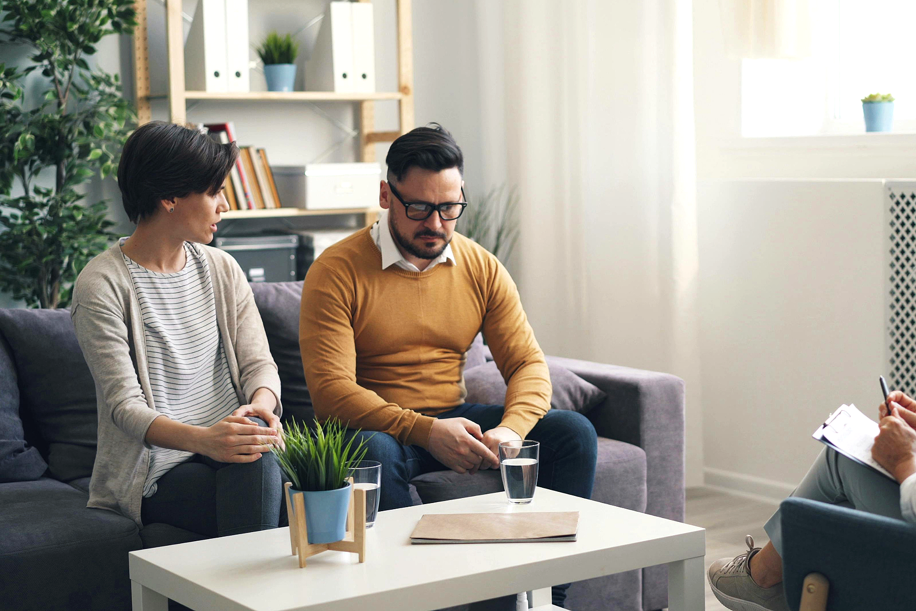 Two people sitting on a gray sofa during a therapy session, with another person taking notes on clipboard in a bright, cozy office with plants and shelves.