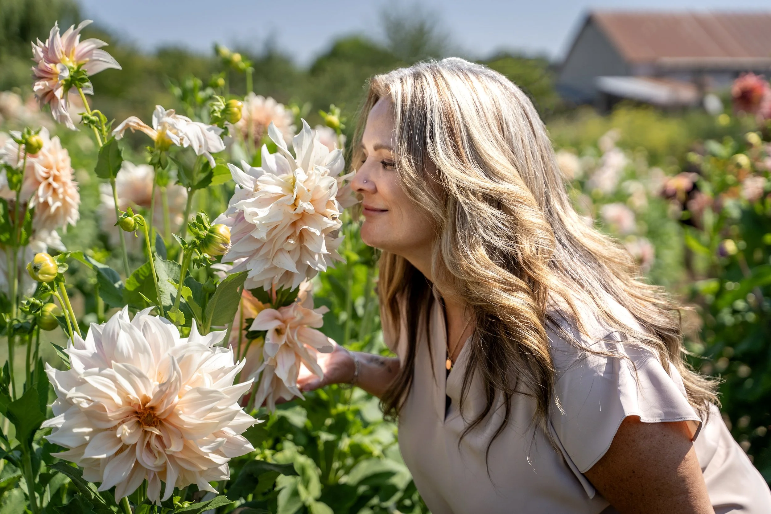 A woman with long, wavy blonde hair and a nose ring smelling pale pink and white dahlias in a garden on a sunny day.