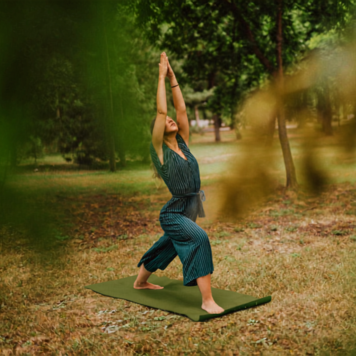 Woman practicing yoga outdoors on a green mat in a park surrounded by trees, performing a pose with hands in prayer position above head.