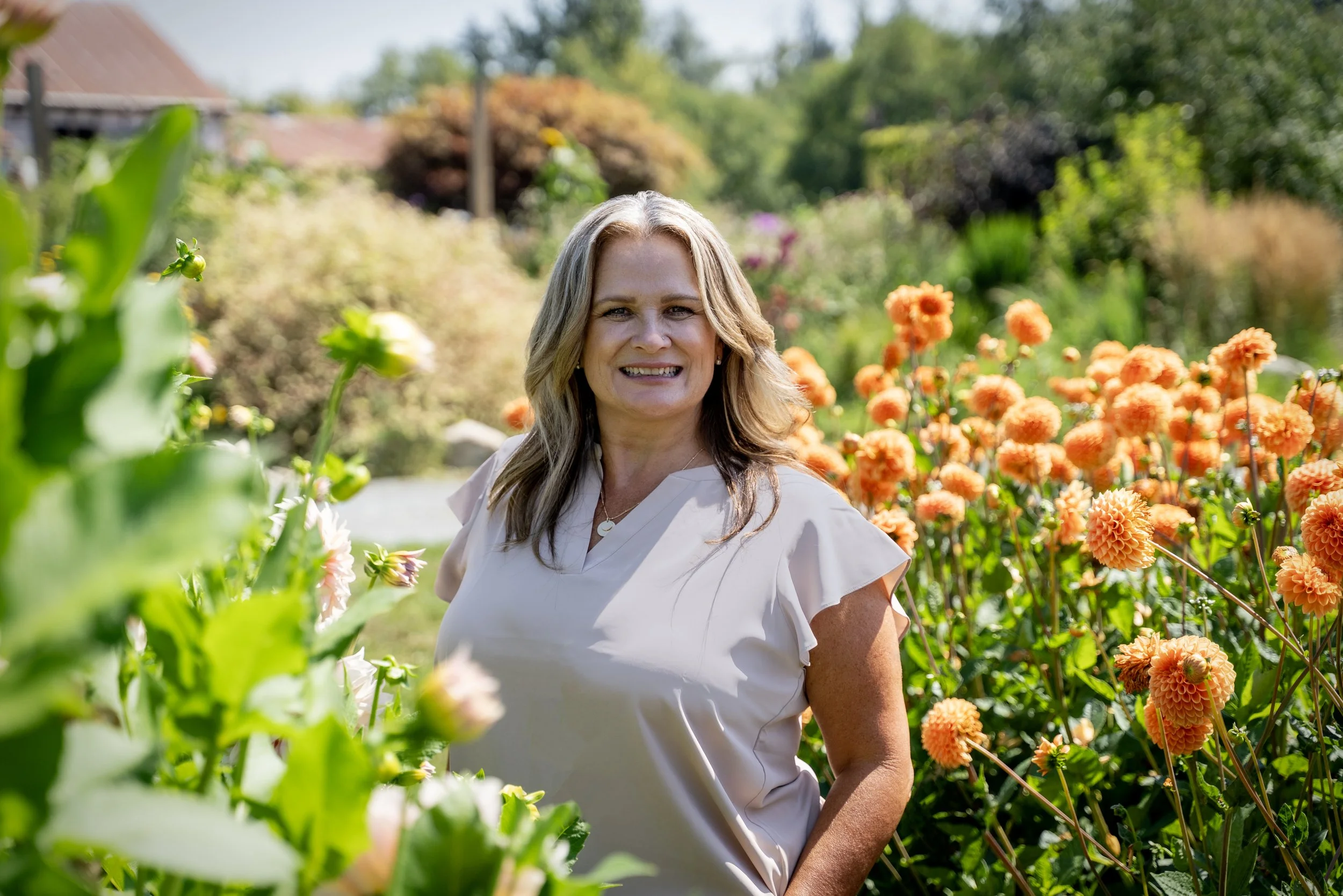 A woman with blonde hair smiling outdoors surrounded by orange and white flowers in a garden.