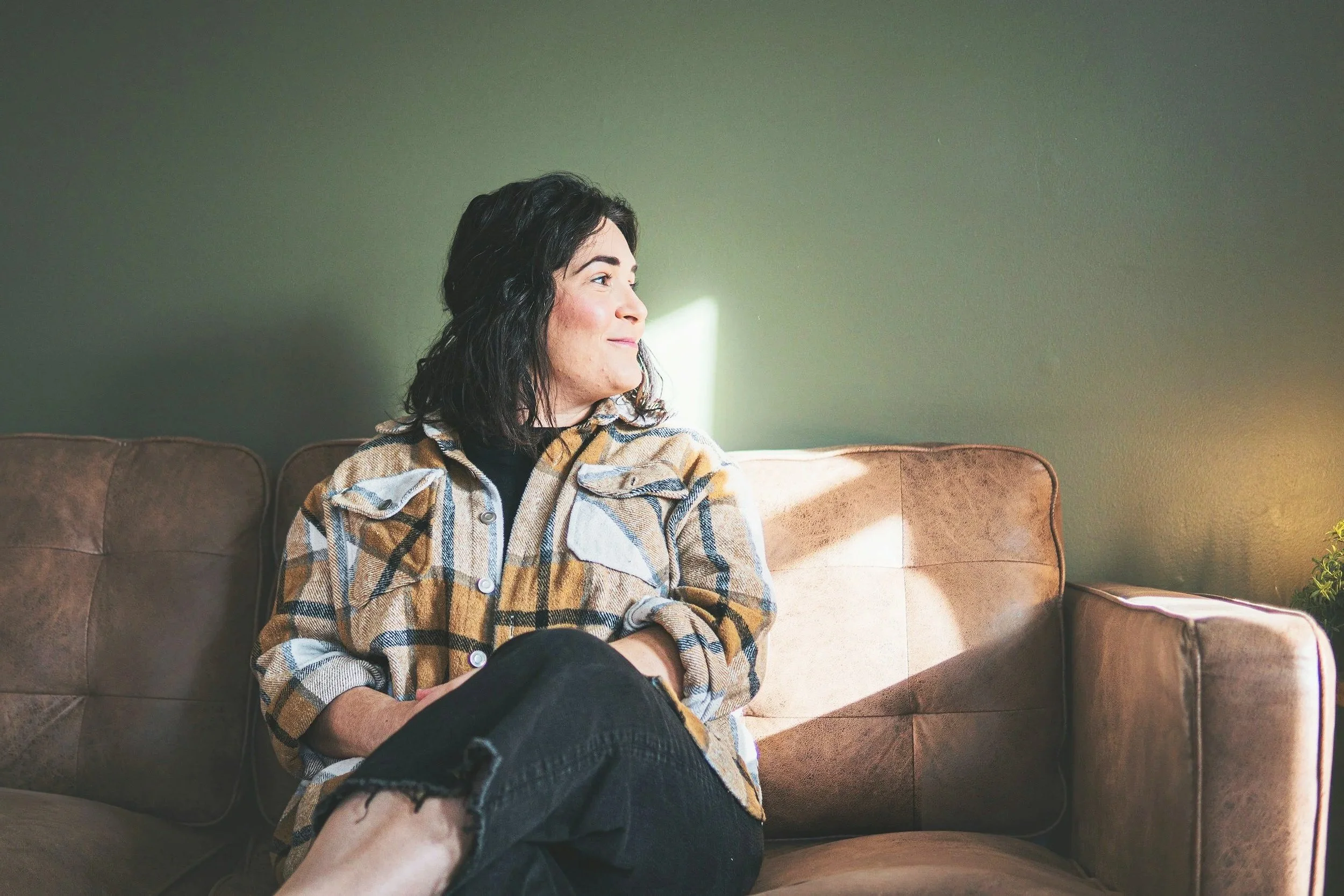 A woman with dark hair and a plaid shirt sitting on a brown couch in a room with green walls, sunlight coming through, looking thoughtfully to the side.