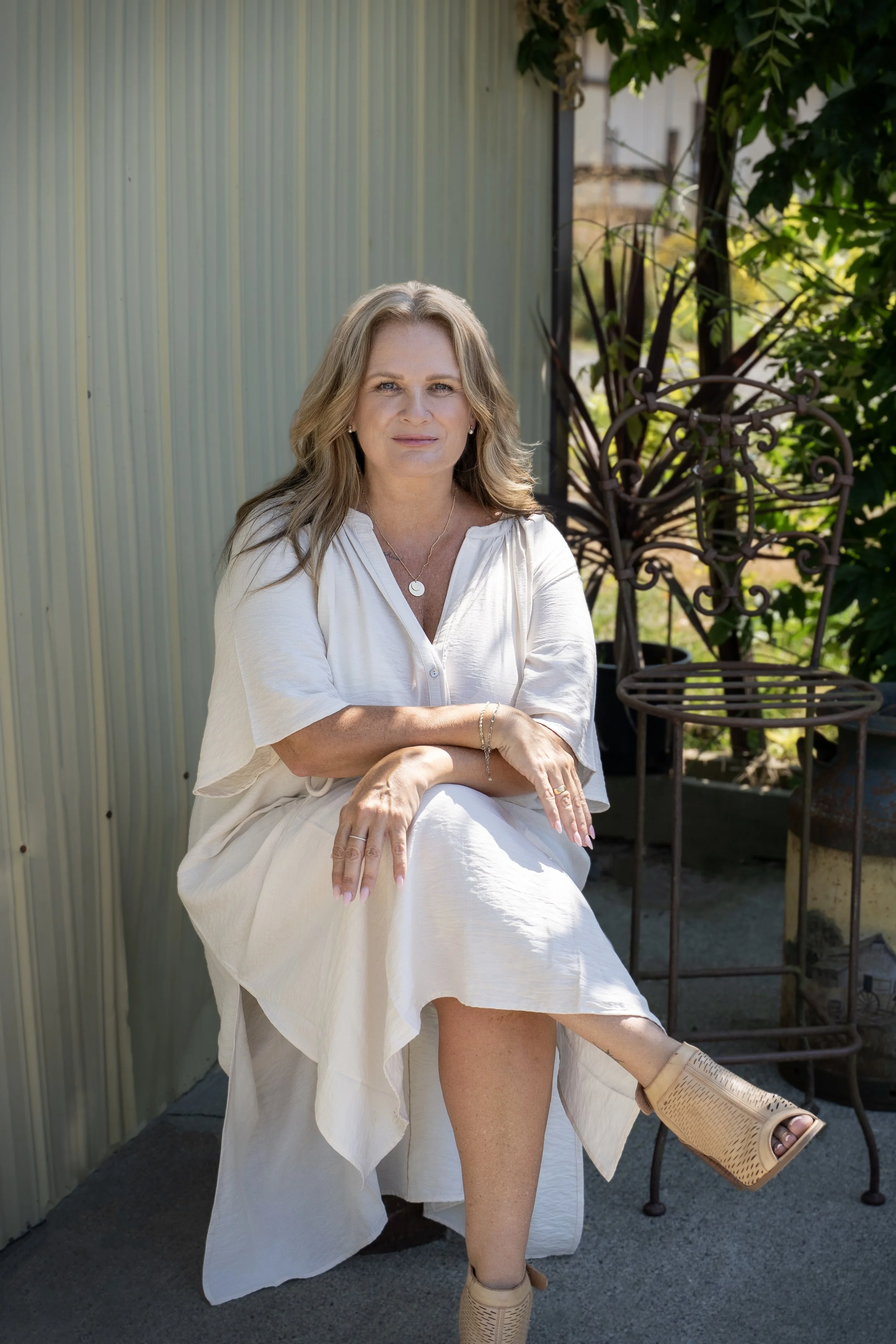 A woman with light brown hair, wearing a white blouse and skirt, sits outdoors with her legs crossed. She is wearing beige open-toe boots, a necklace, and a bracelet, and has a slight smile. Behind her are green plants and a decorative metal chair.